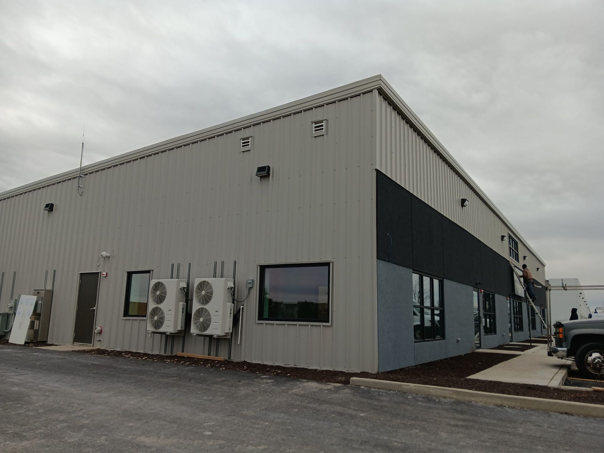 Gray industrial building with black accents and air conditioning units under an overcast sky.