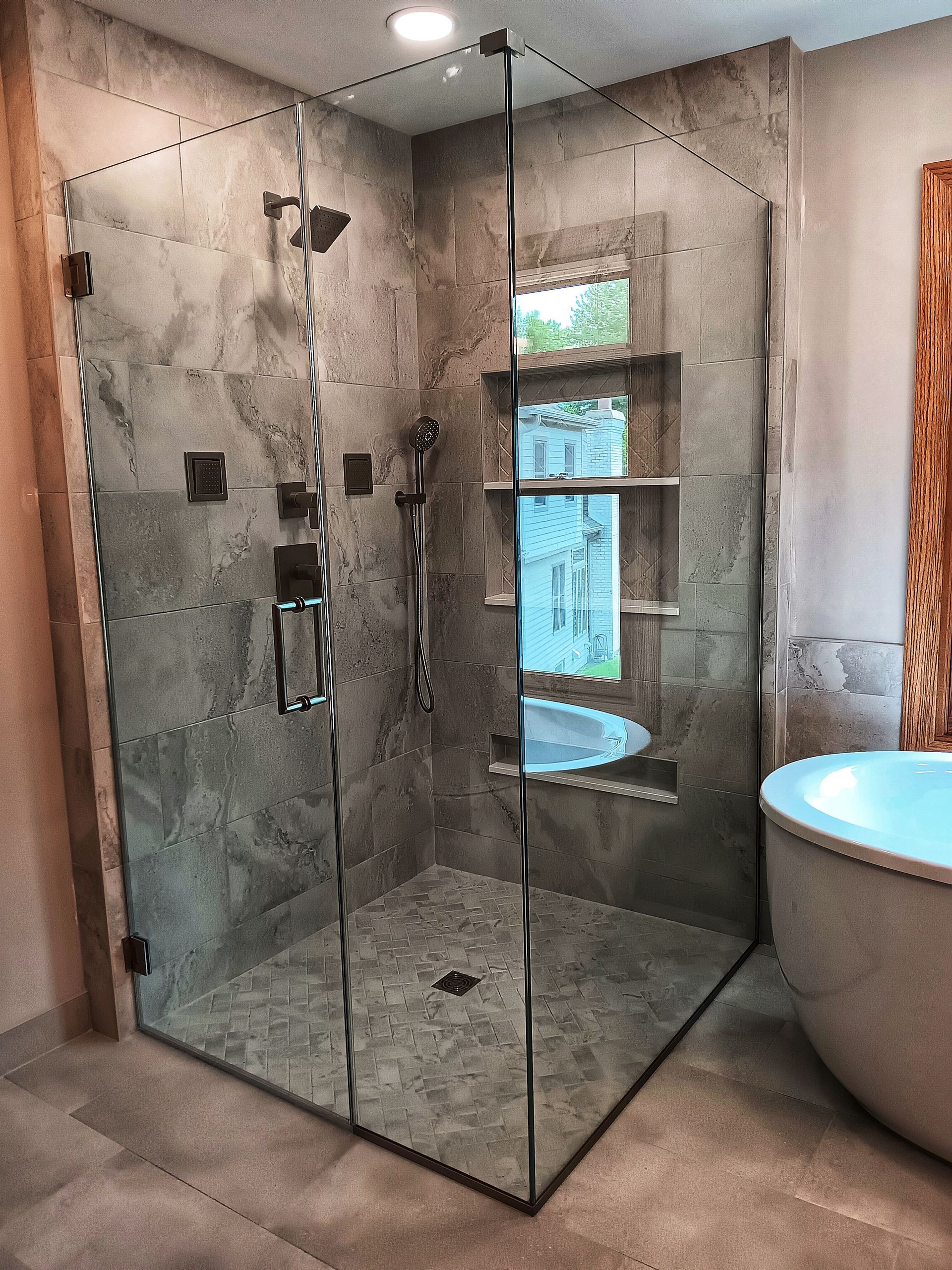 Modern, gray-tiled shower with glass doors and fixtures. Adjacent is a white tub and open window.