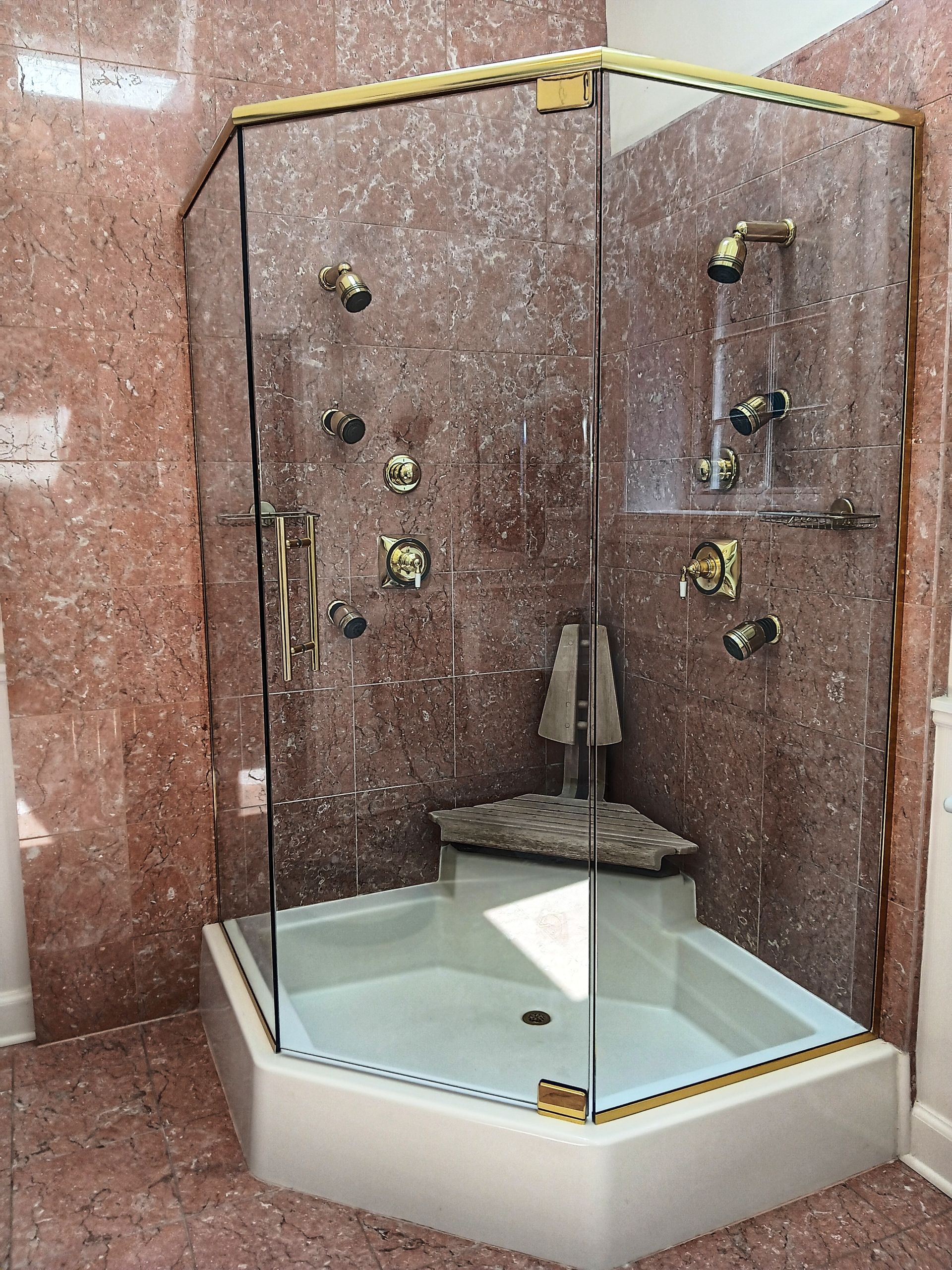 A luxurious shower with glass doors and gold fixtures against a reddish-brown marble wall.