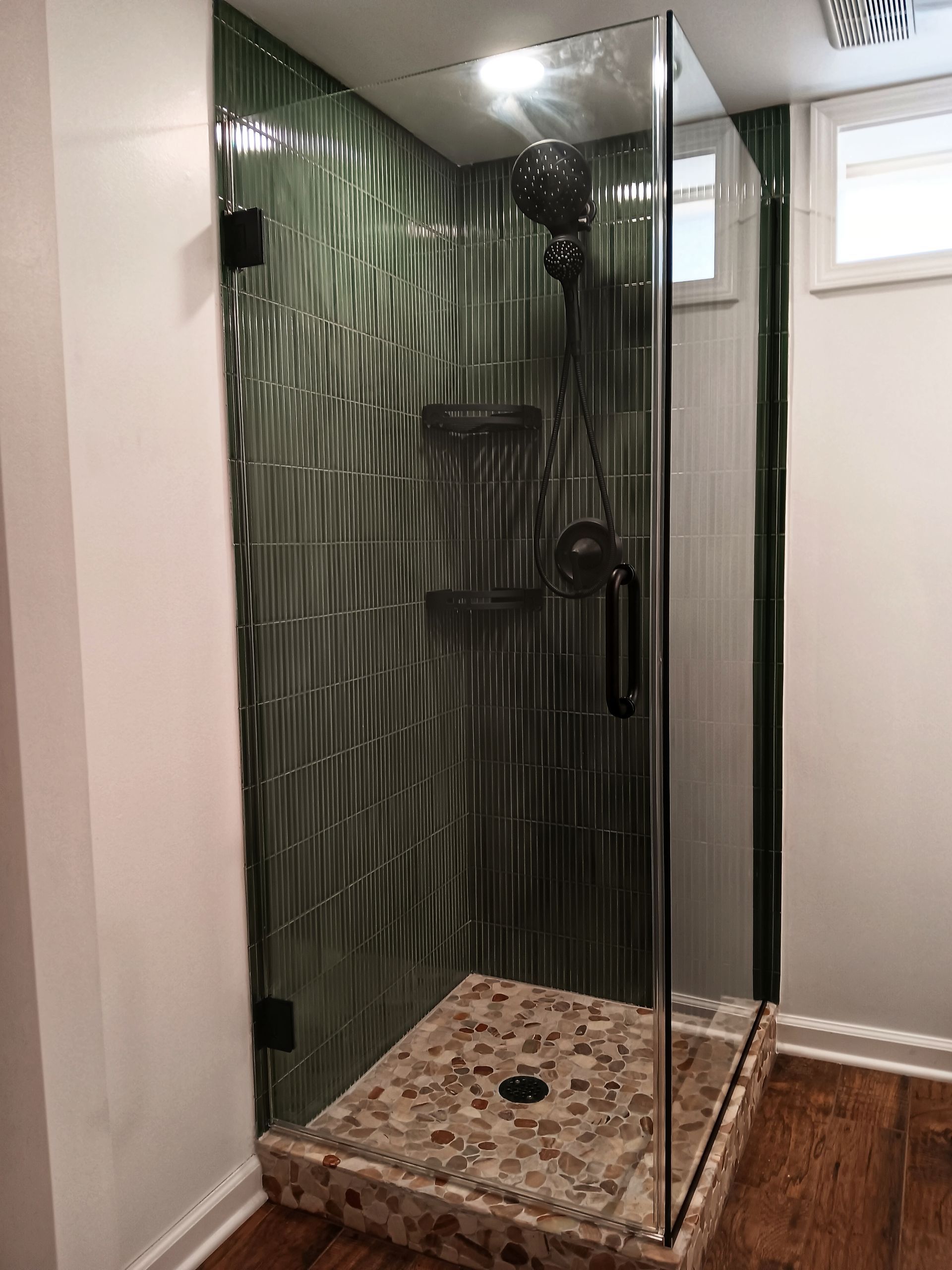 Green-tiled shower with glass door and stone floor in a corner of a room with wooden flooring and white walls.