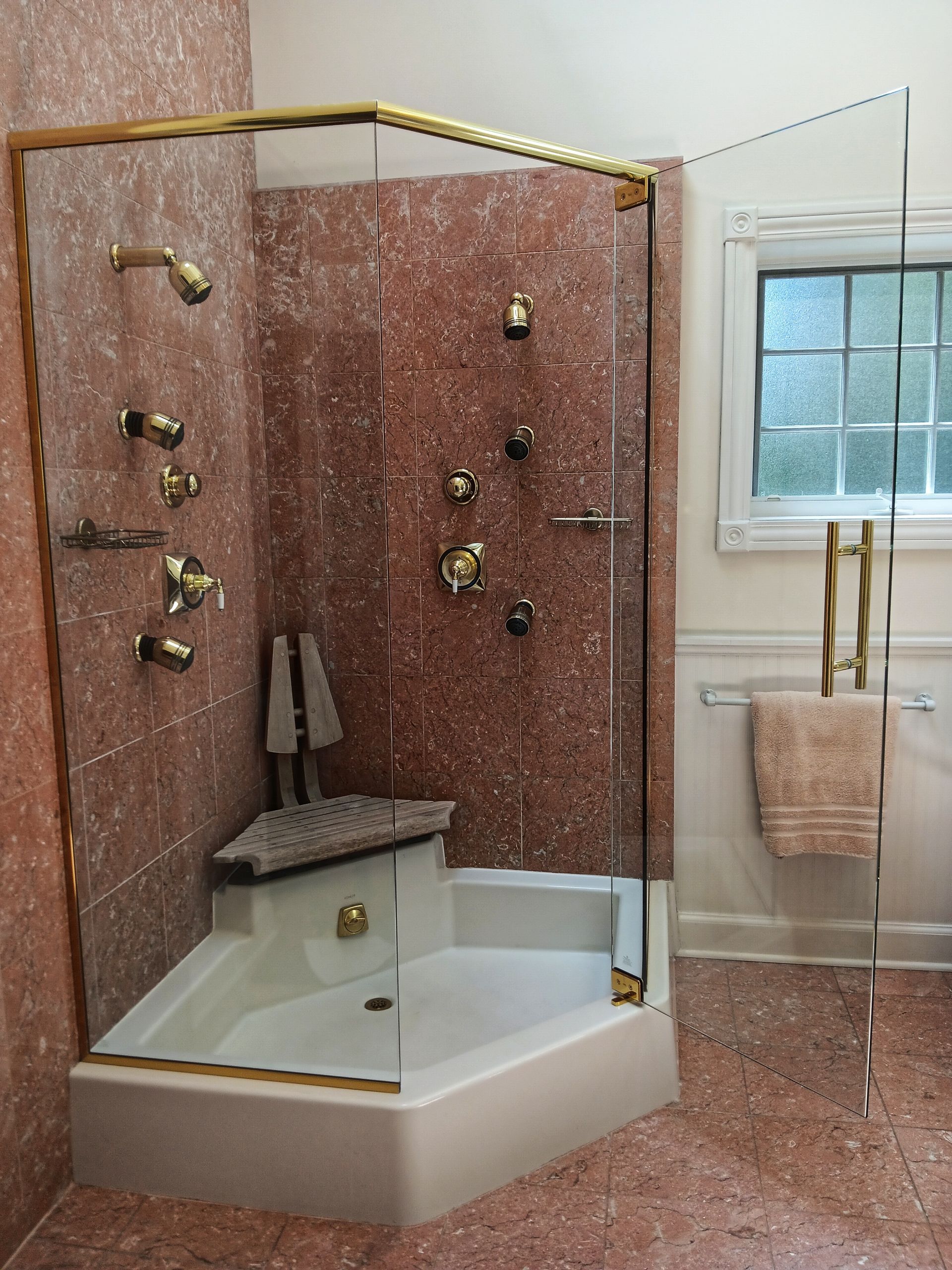 Corner shower with gold trim, red speckled walls, white base, seat, and multiple gold showerheads.
