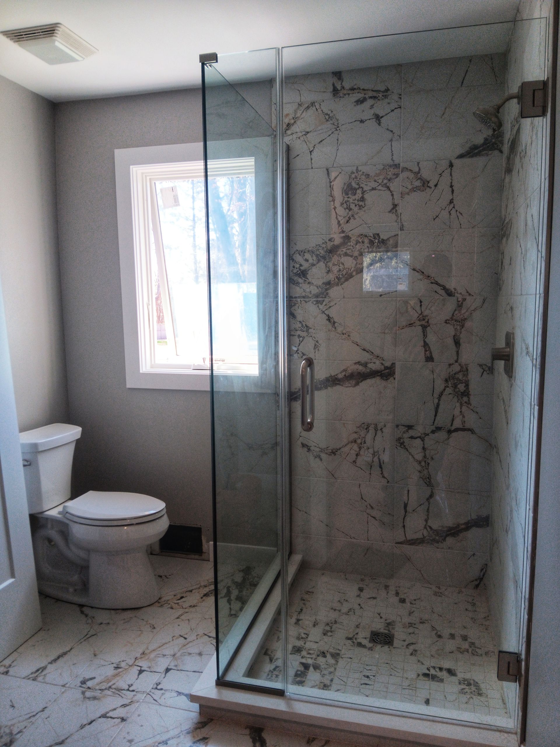 Bathroom with a glass shower, toilet, window, and marble-like tile.