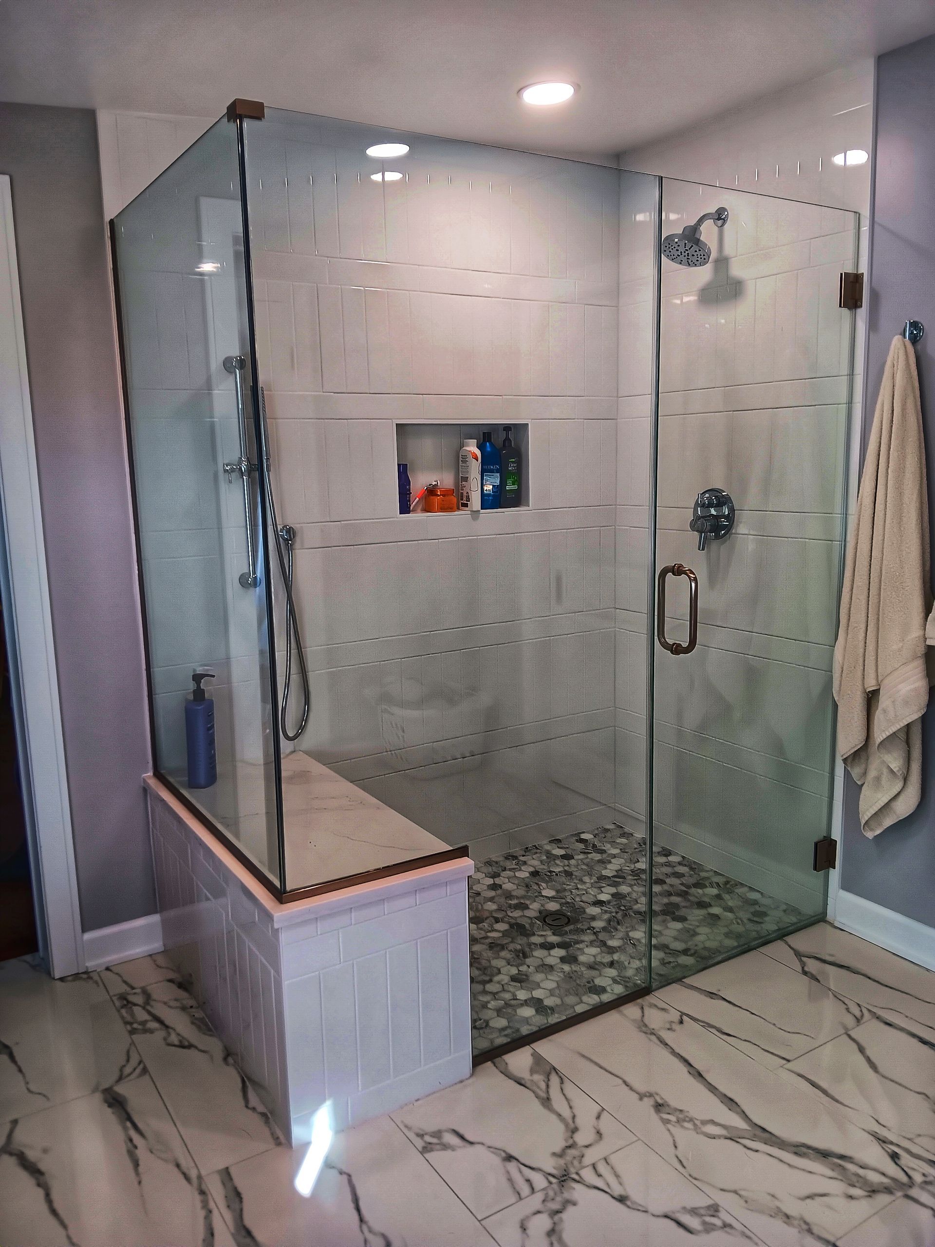 A modern shower with glass walls, bench, and stone floor.  White and gray tile, and chrome fixtures.