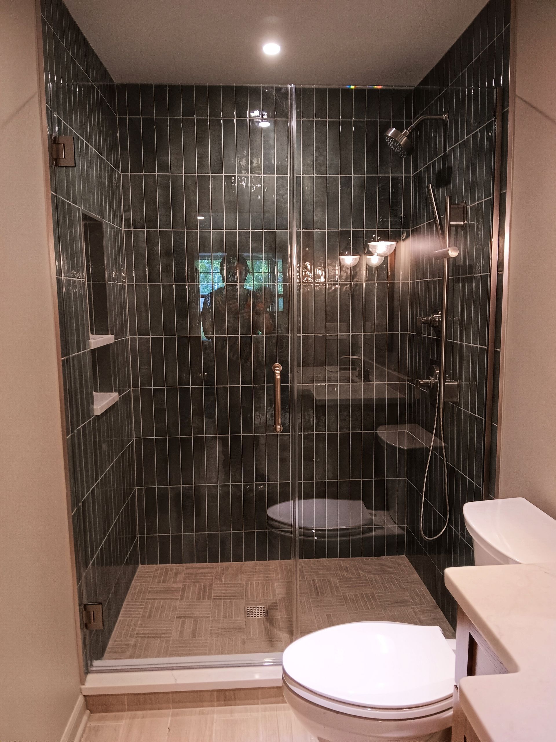 Shower with dark tile walls, glass door, toilet, and light-colored floor and walls.