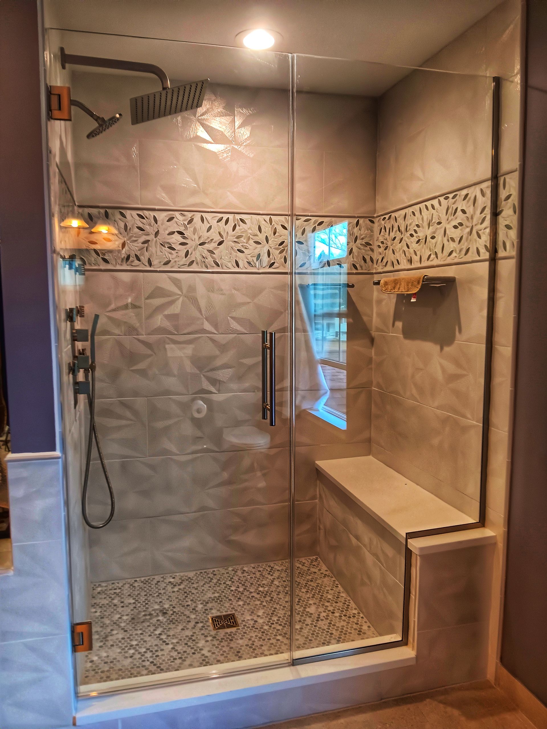 Modern bathroom shower with glass door and bench seat, gray and beige tiles, and pebble floor.