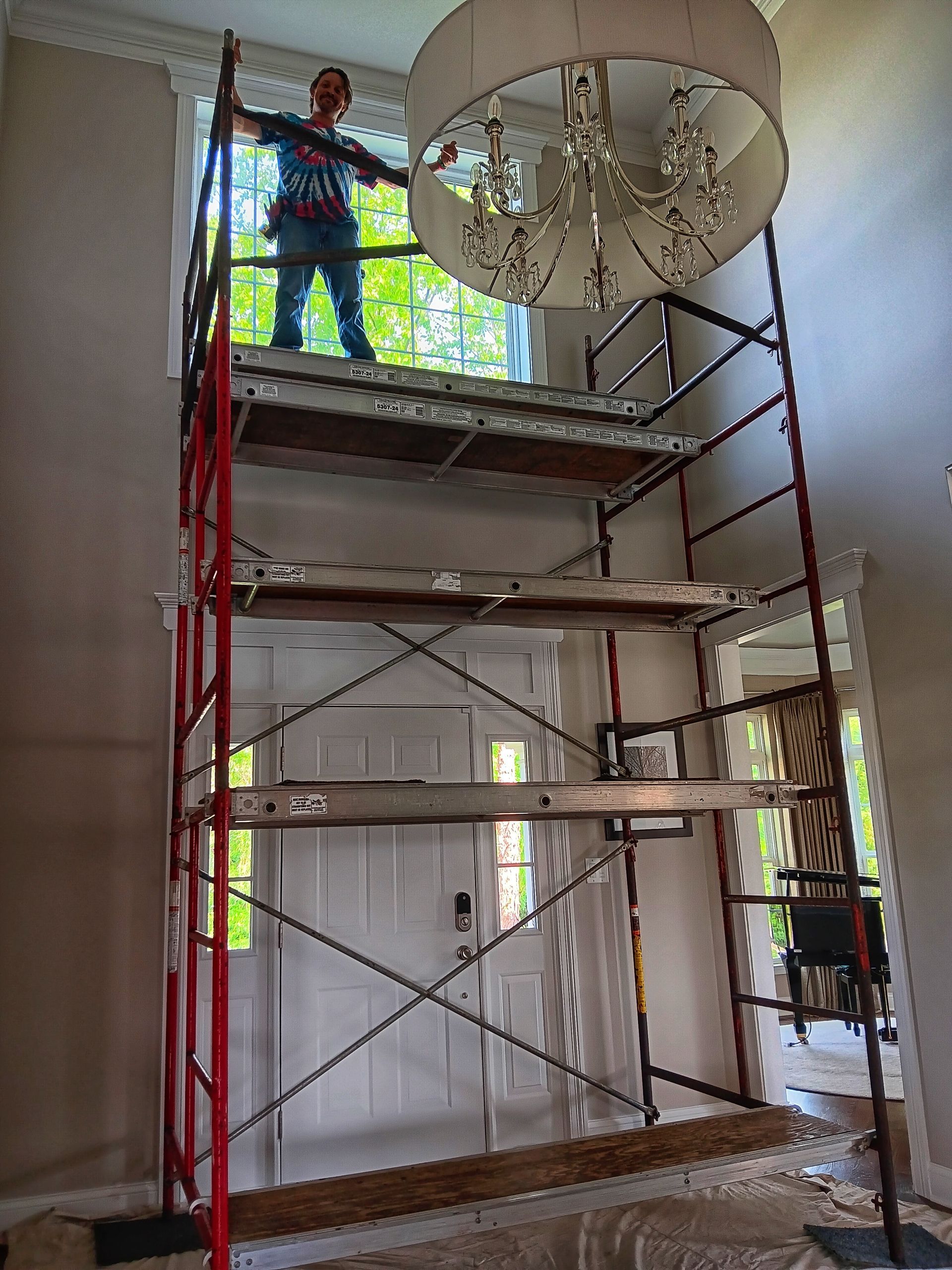 A person on a tall scaffold near a window and chandelier inside a home.