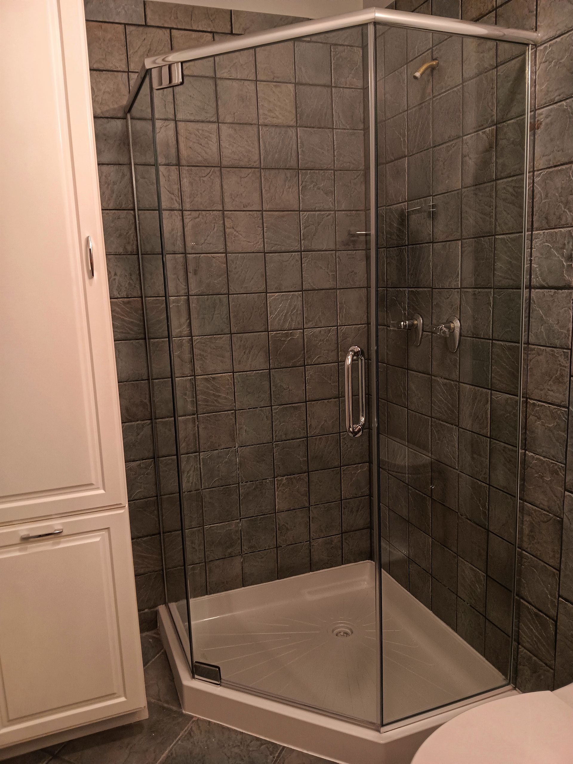 A corner shower with clear glass doors and textured gray tile walls, next to a white cabinet.