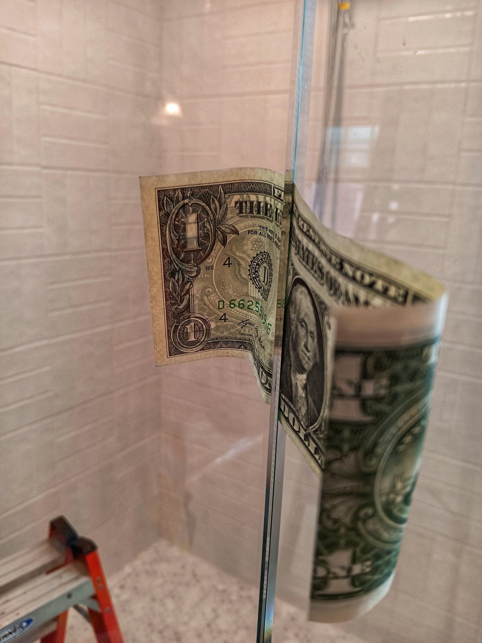 A dollar bill is stuck to a glass shower door. The bill is slightly rolled.