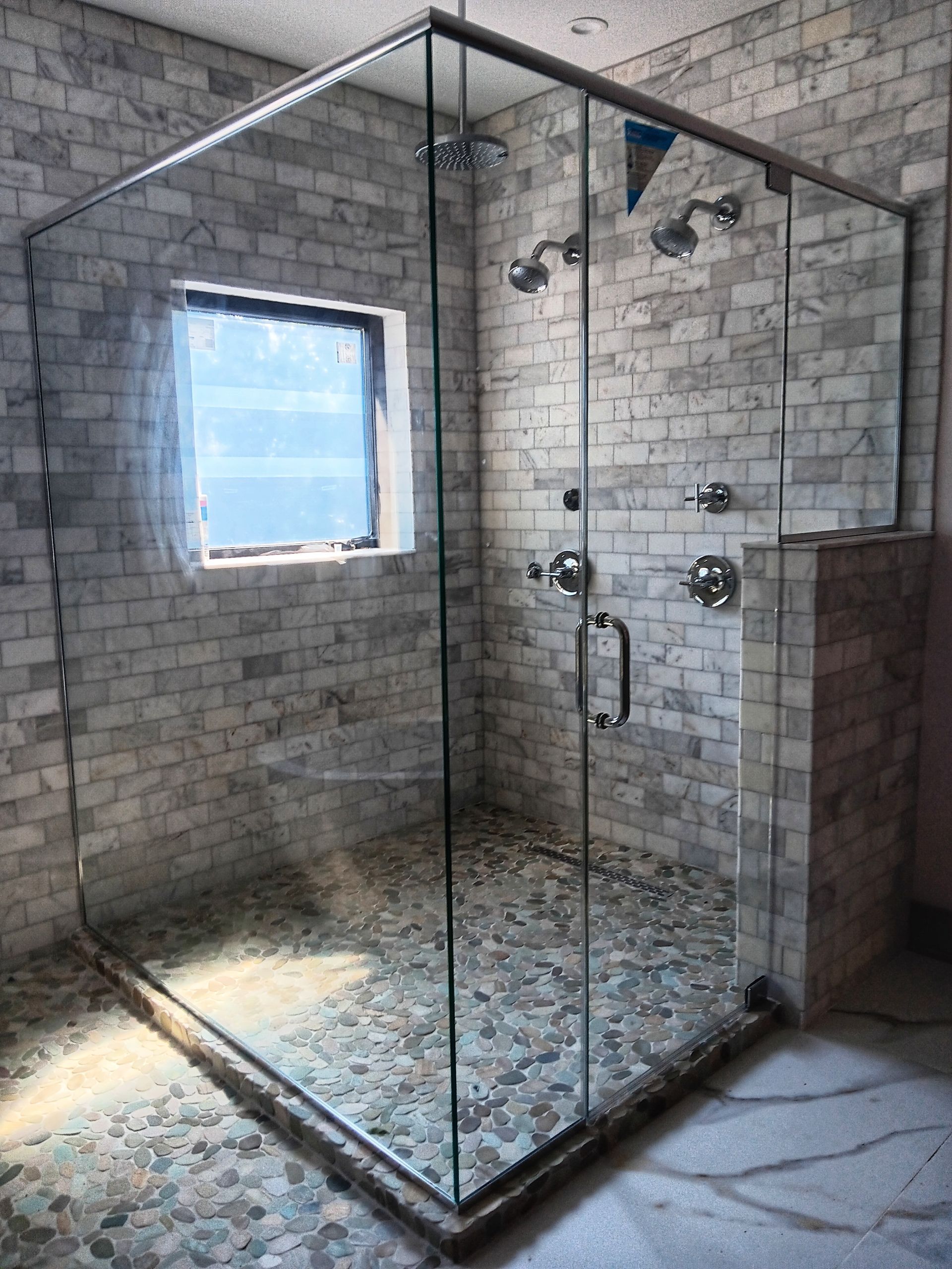 Glass-enclosed shower with pebble floor and brick-style tiled walls; window on one side, multiple showerheads.