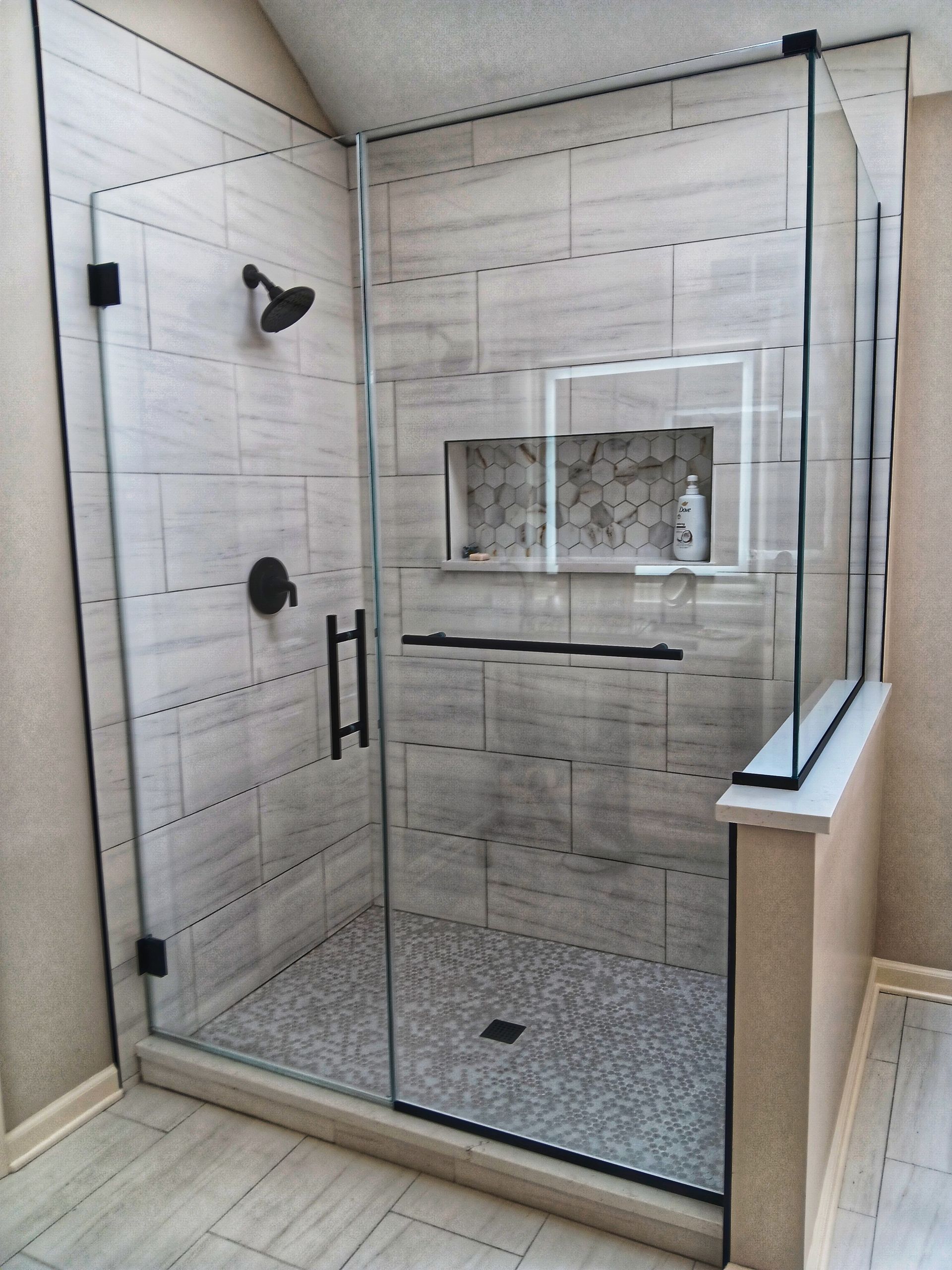 Glass-enclosed shower with tiled walls and pebble floor.  Black hardware. Beige tile and walls.