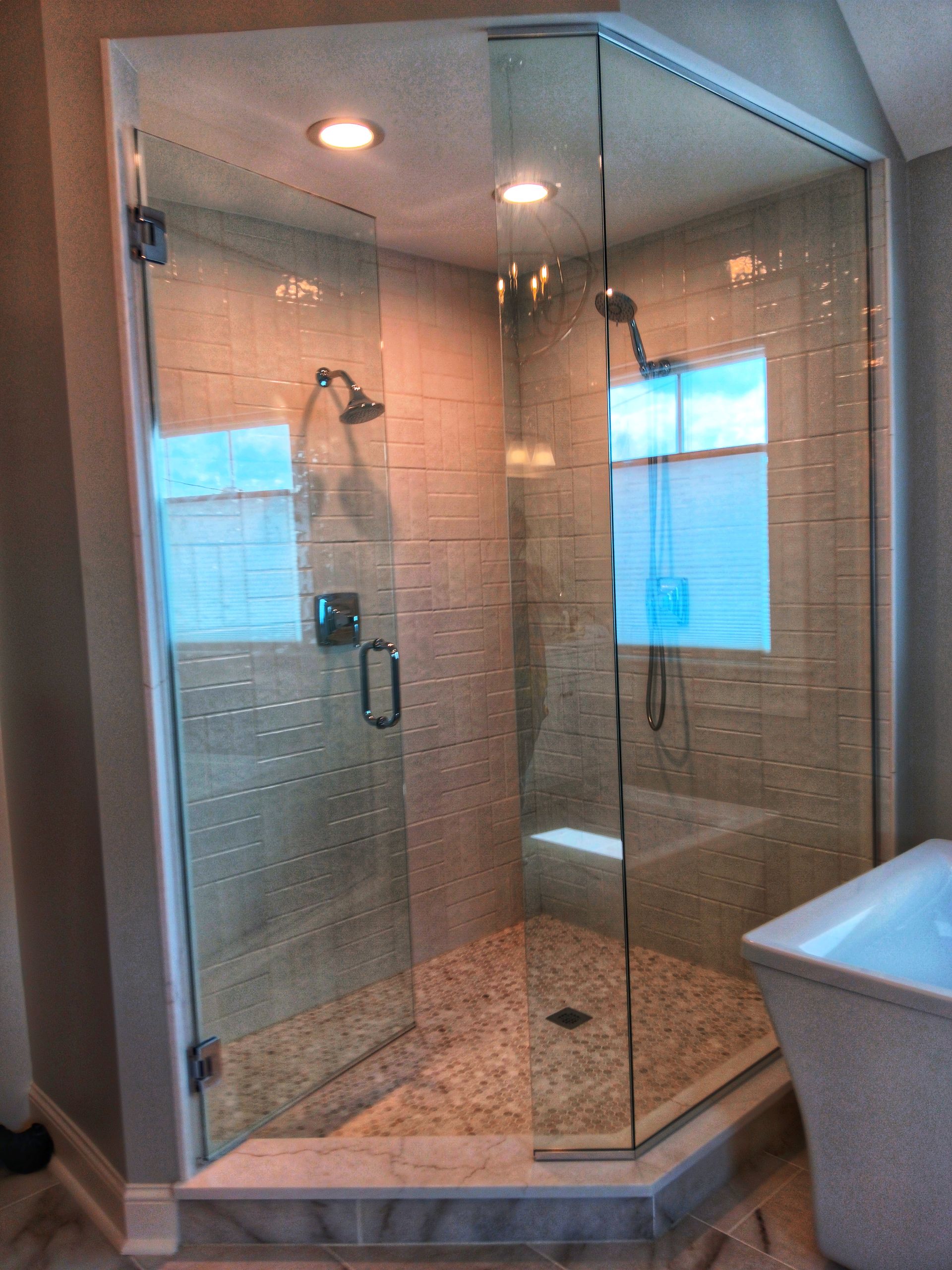 Glass-enclosed corner shower with glass door and rain shower head. Stone tiled floor and beige walls.
