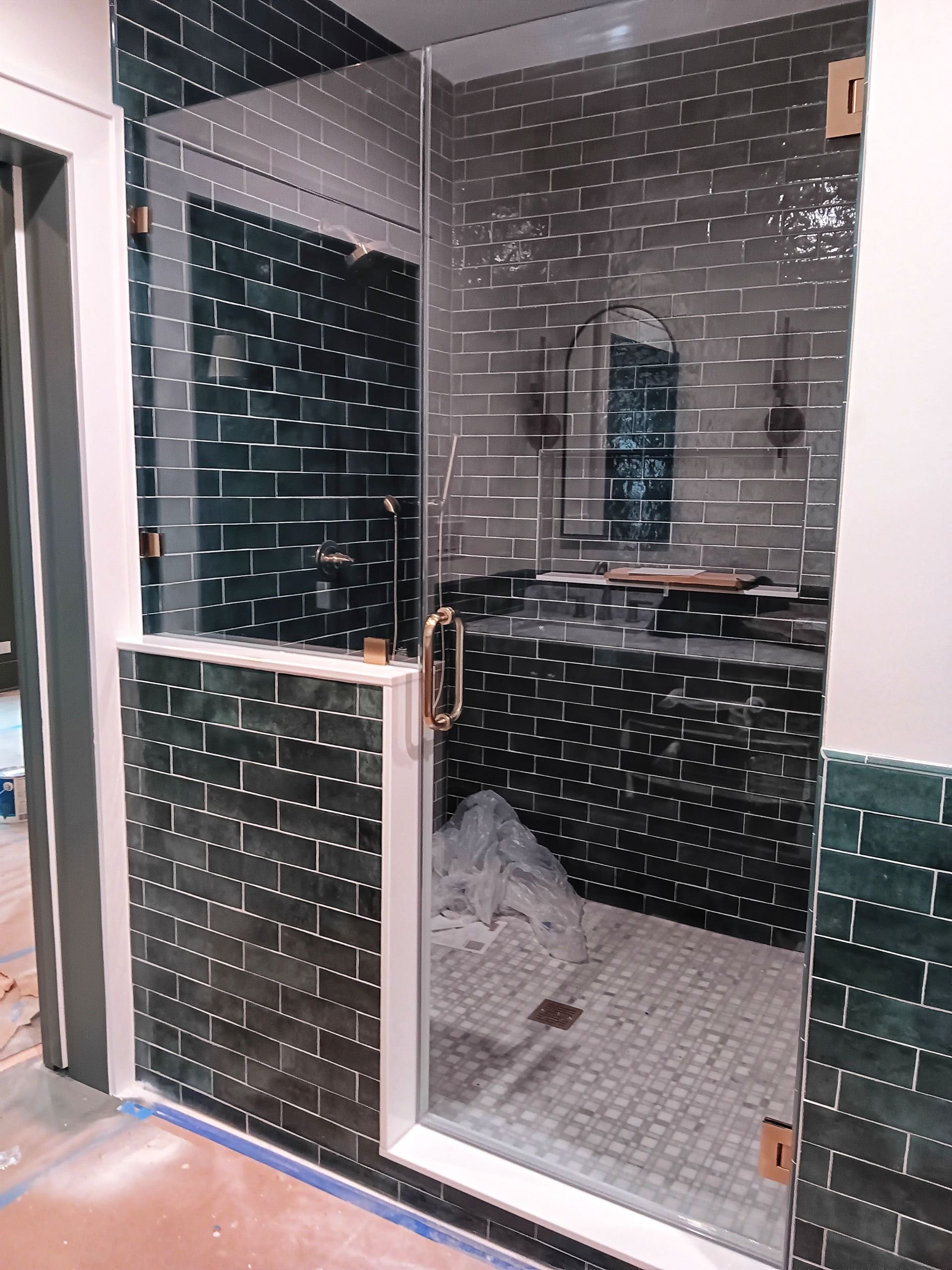 A walk-in shower with dark green tiled walls and glass door; gold hardware; white and silver tiled floor.