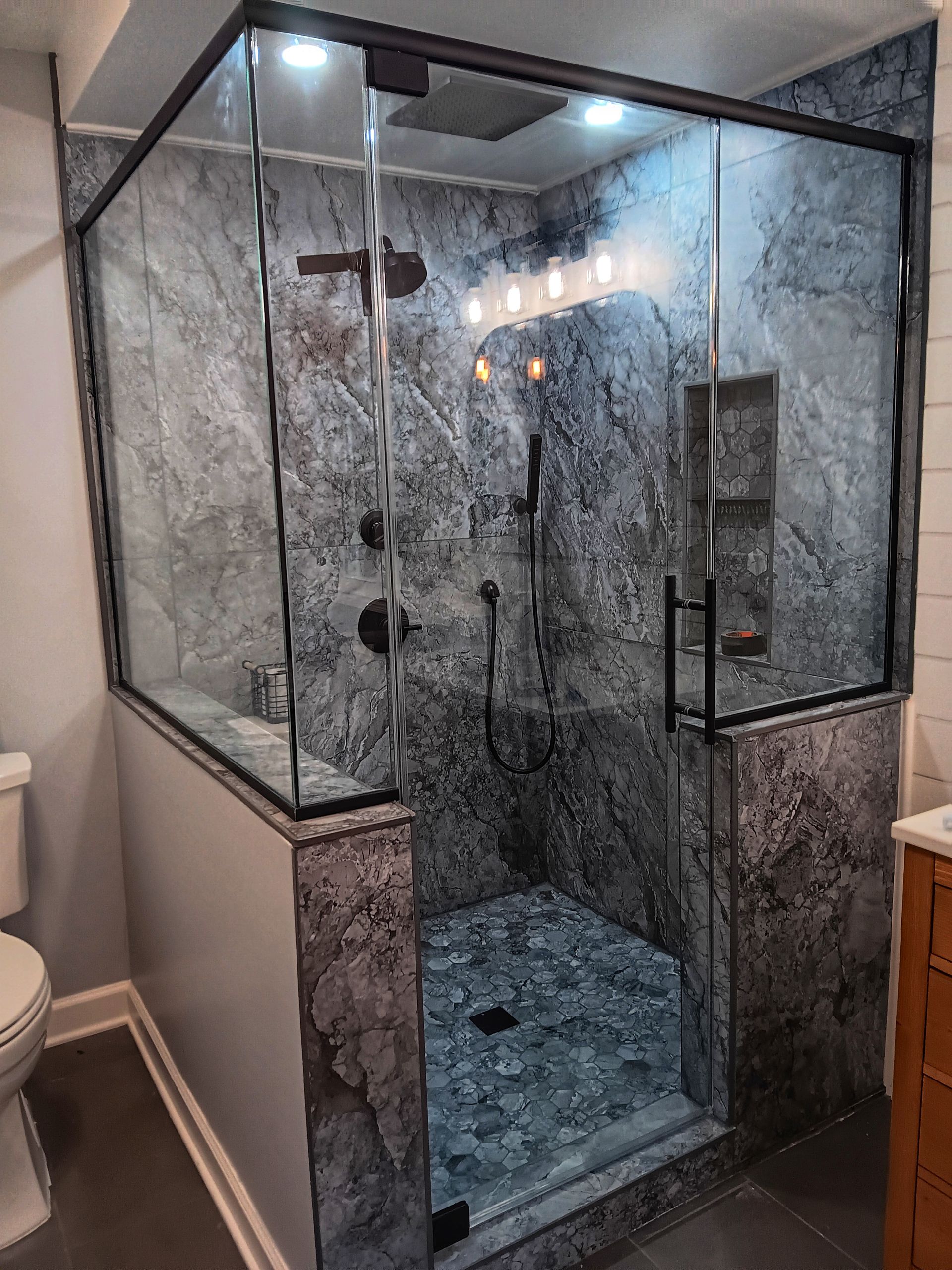 Modern shower with gray stone-like walls, glass door, black fixtures, and pebble tile floor.