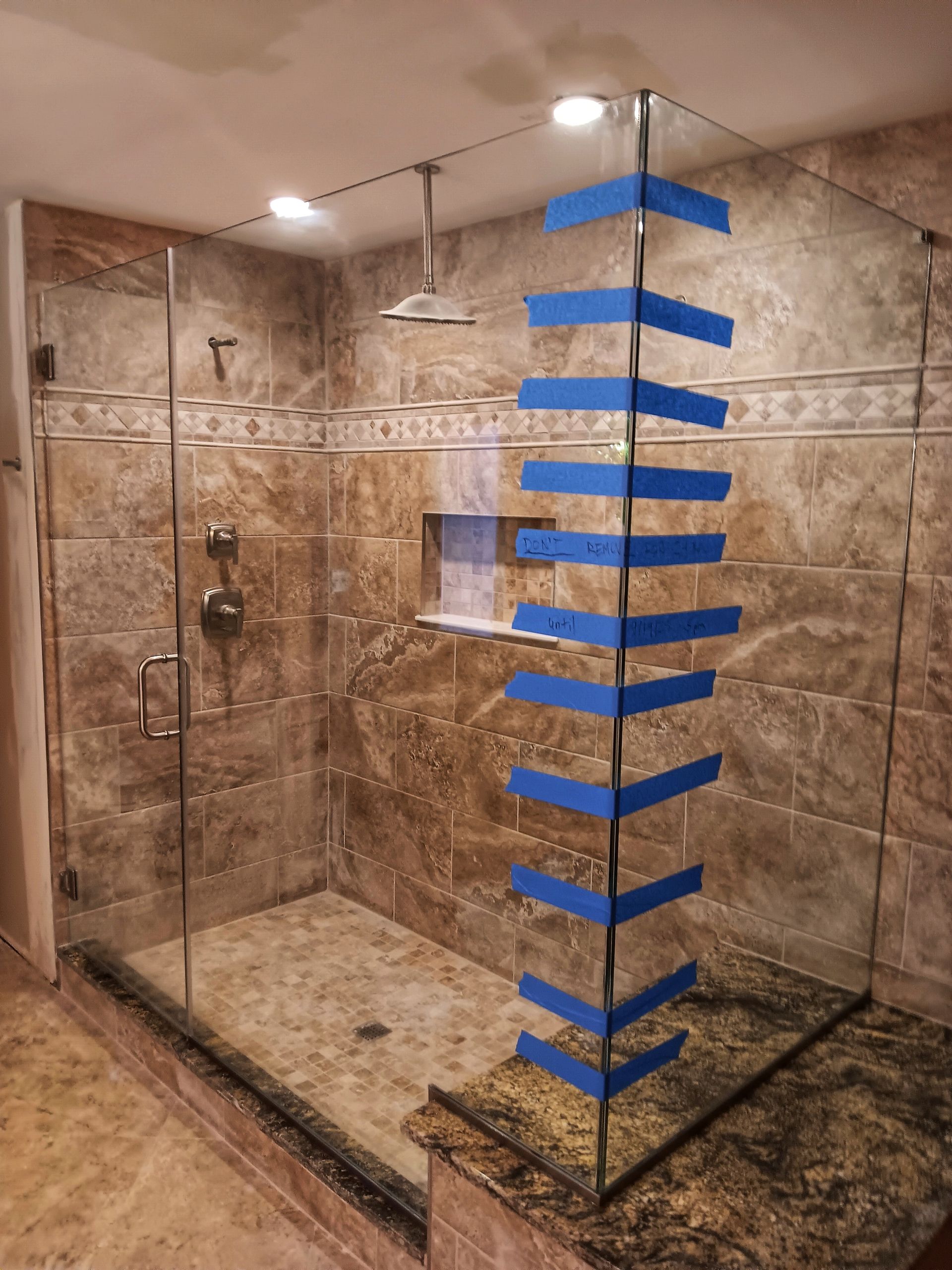 Shower with glass walls, brown tile, and a granite bench. Blue tape marks the corner.