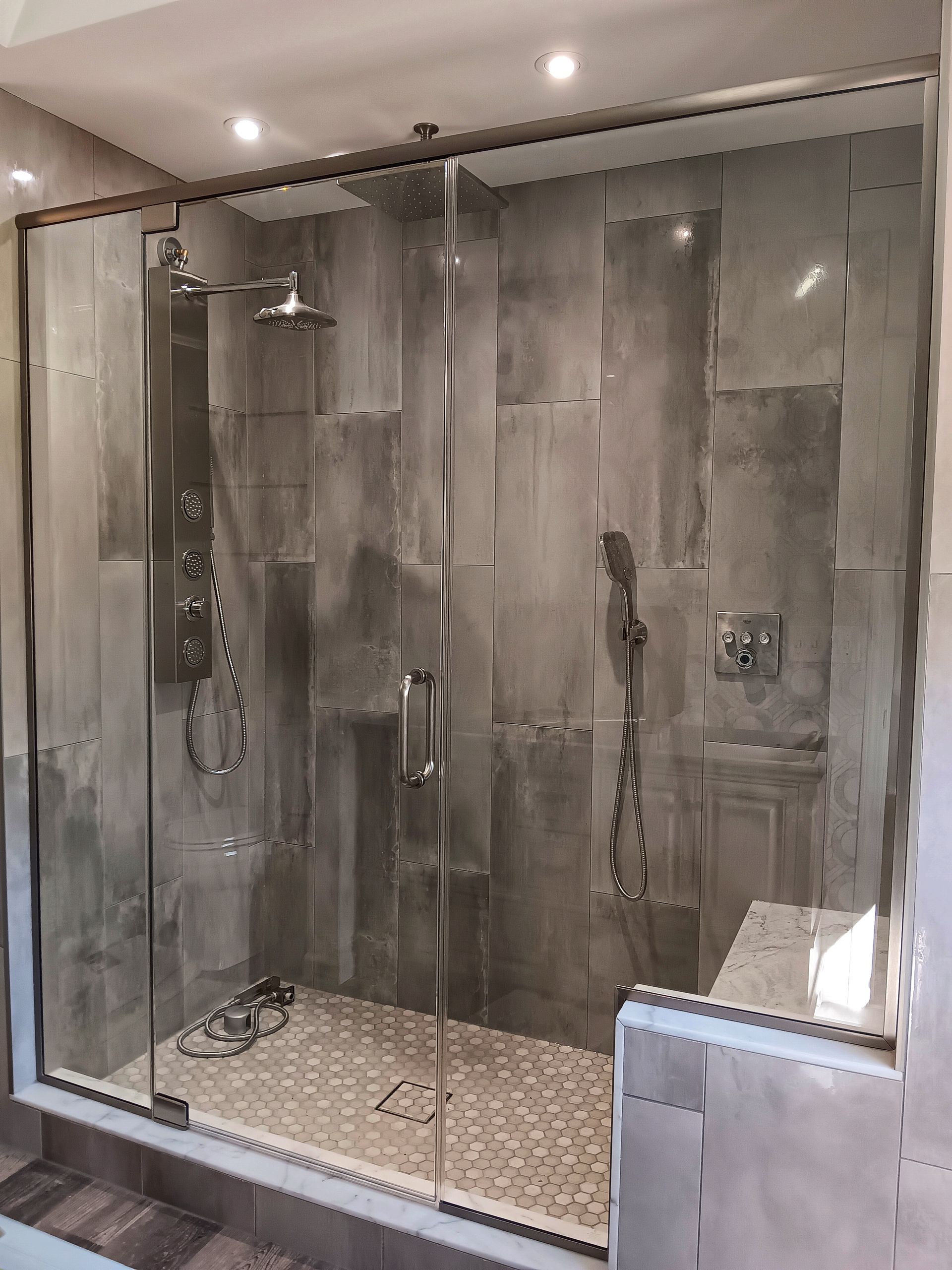 Modern gray tiled shower with glass doors, two shower heads, and a built-in bench.