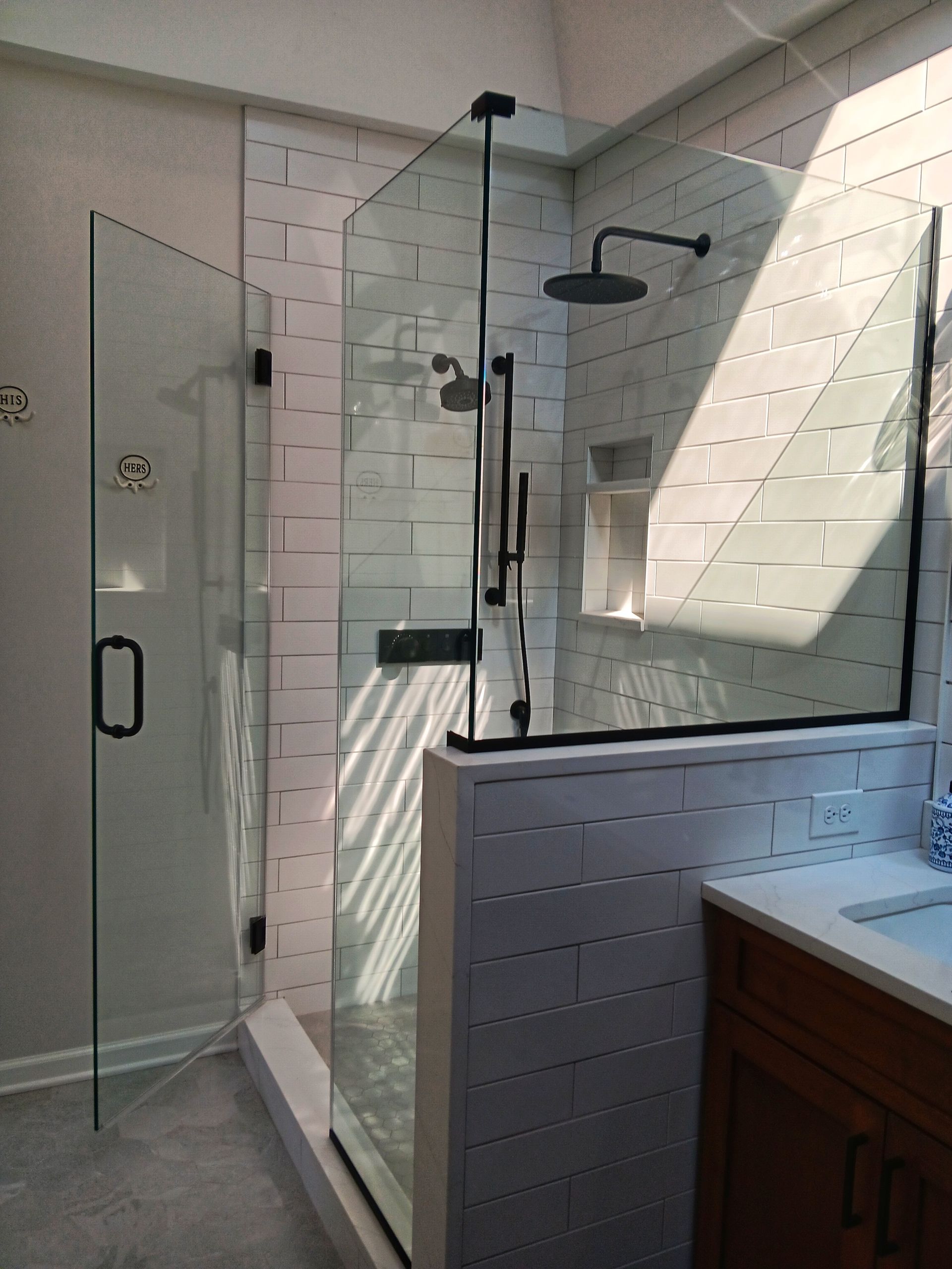 Glass-enclosed shower with black fixtures and white tiled walls. A part of a vanity is visible.