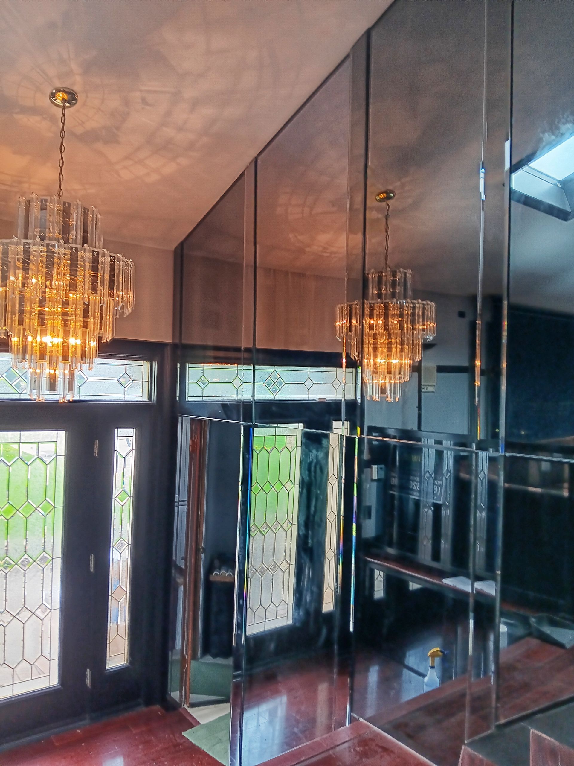 Entryway with mirrored walls, chandeliers, and a black front door. Reflective surfaces create depth and light.