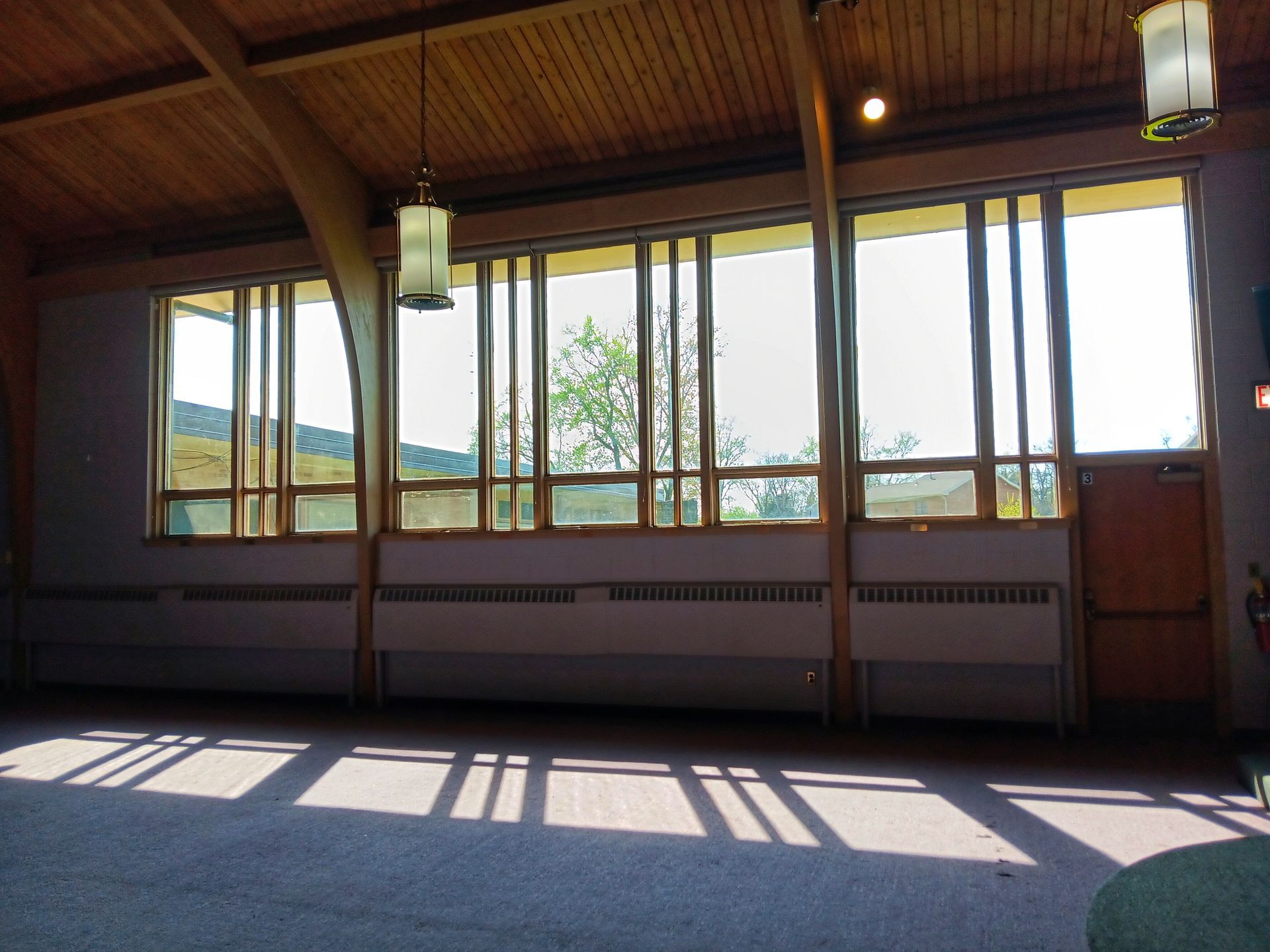 Interior view with large windows, light streaming in. Wooden beams, pink carpet, and radiator below the windows.
