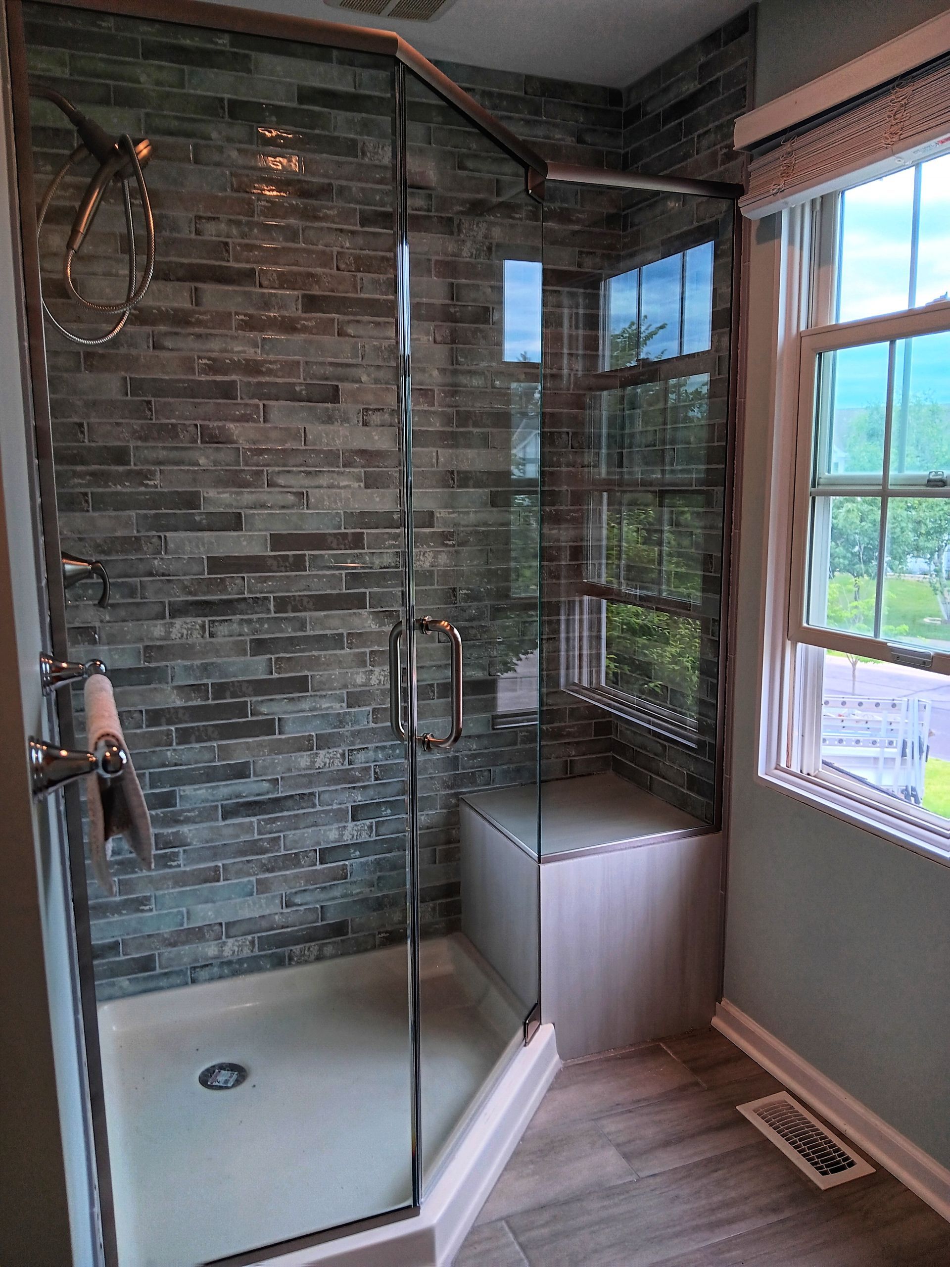 Corner shower with glass doors, built-in seat, grey tile wall, and a window to the right.