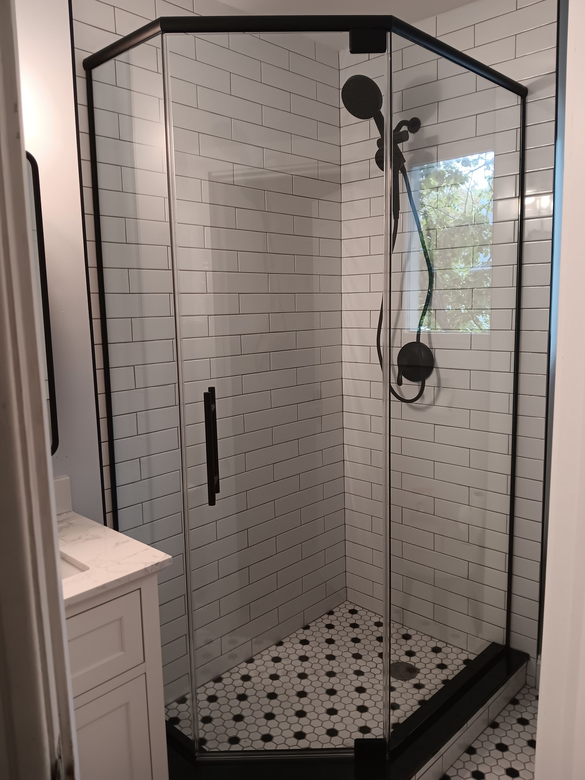 Black-framed glass shower in a bathroom with white brick-style tile and black and white patterned floor tiles.