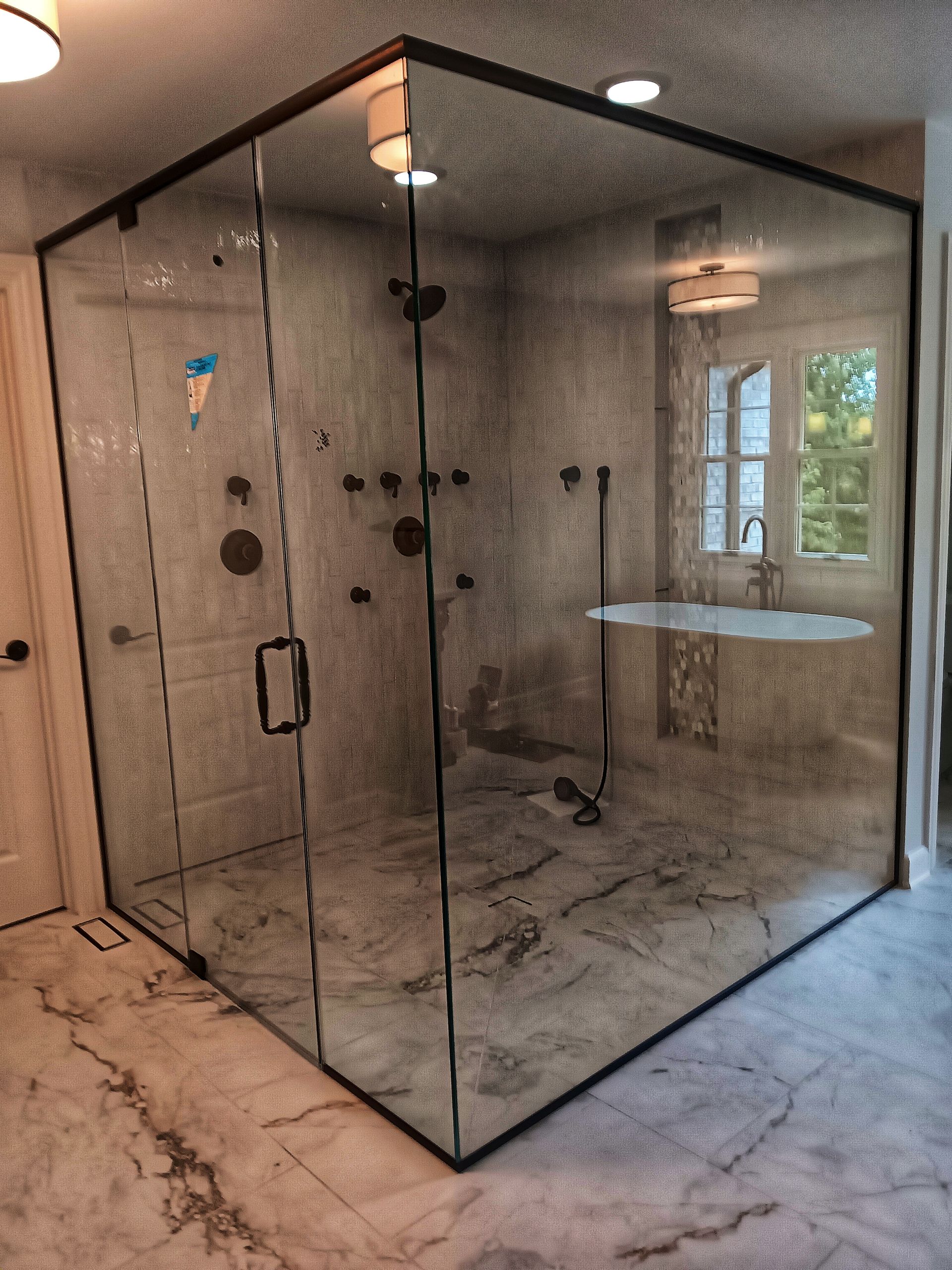 Spacious, glass-enclosed shower with black trim. Features multiple showerheads and a marble tile floor, next to a window.