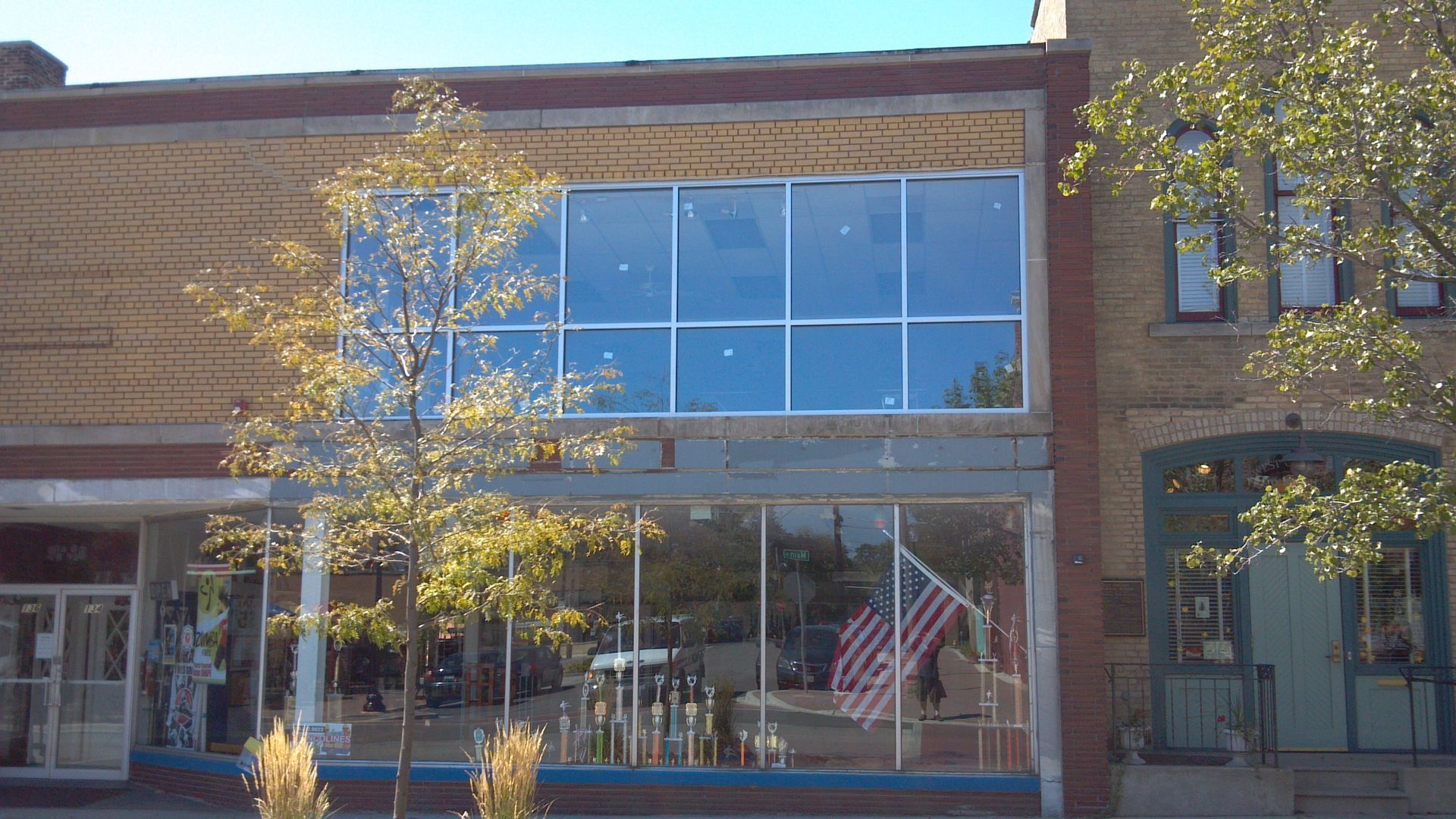 Storefront with large windows, American flag, brick facade, trees, sunny day.