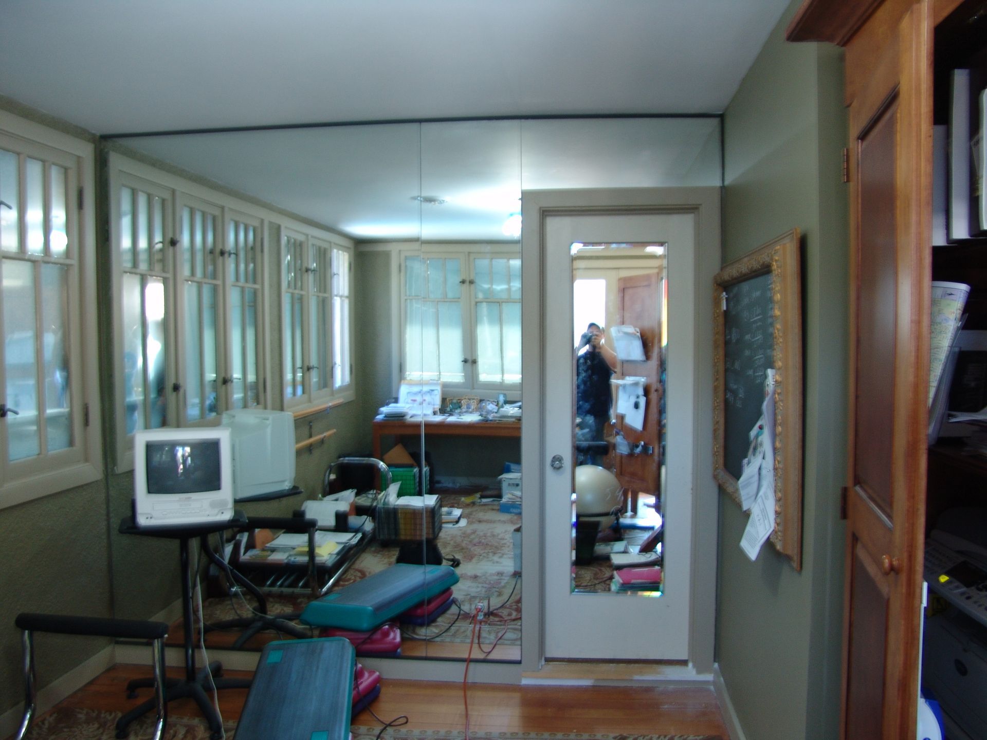 Room with mirror wall, desk, windowed wall, and a doorway with a mirrored door, appearing cluttered.
