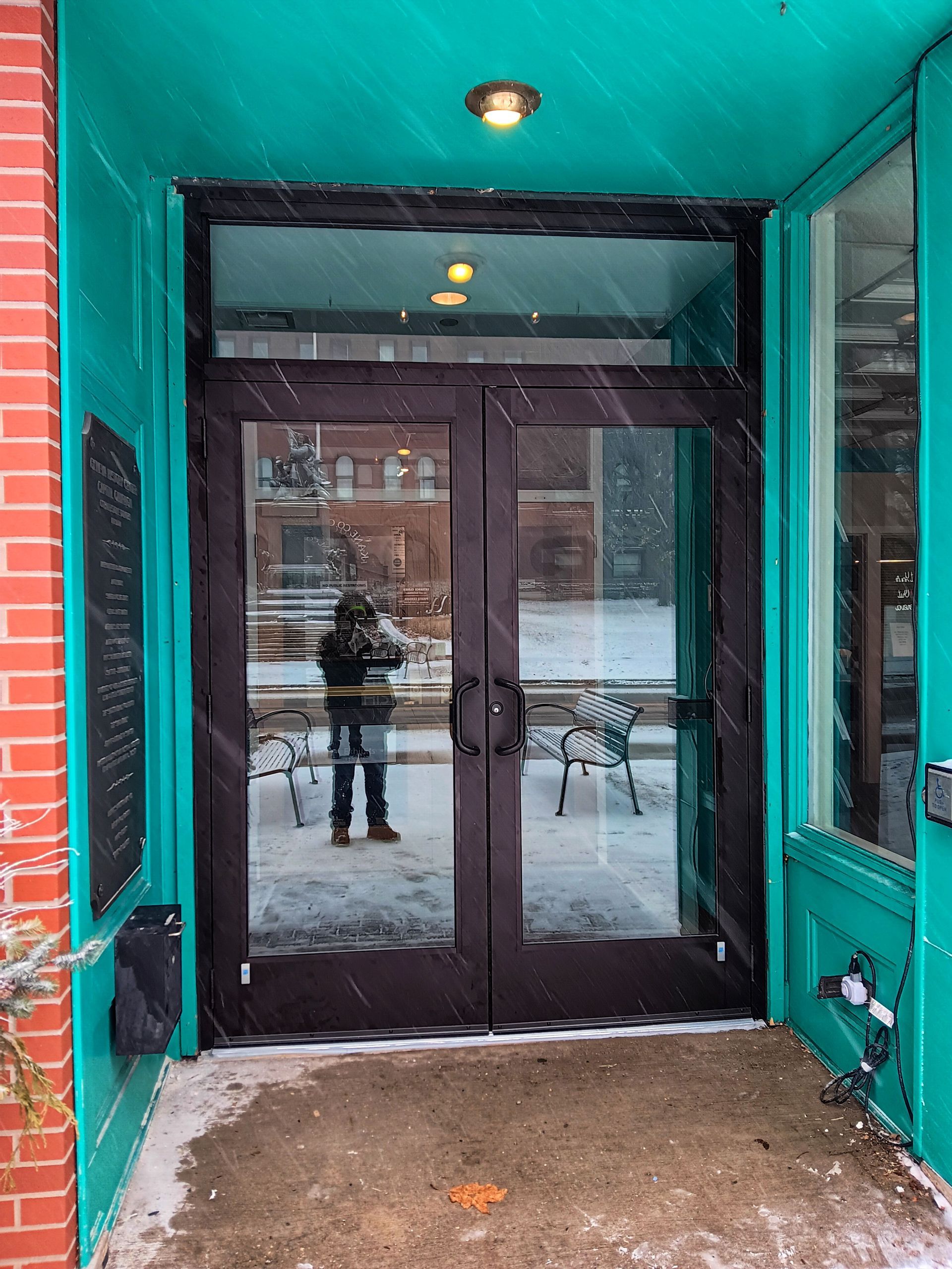 Double glass doors with a person reflected in the glass, teal frame, snowing outside.