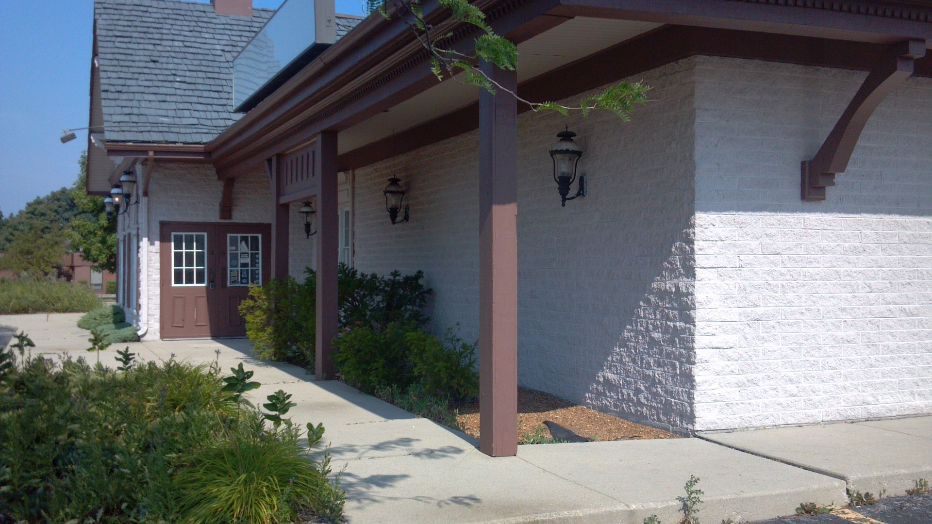 Building exterior with a brown awning, columns, and door. Cream-colored walls and green bushes.
