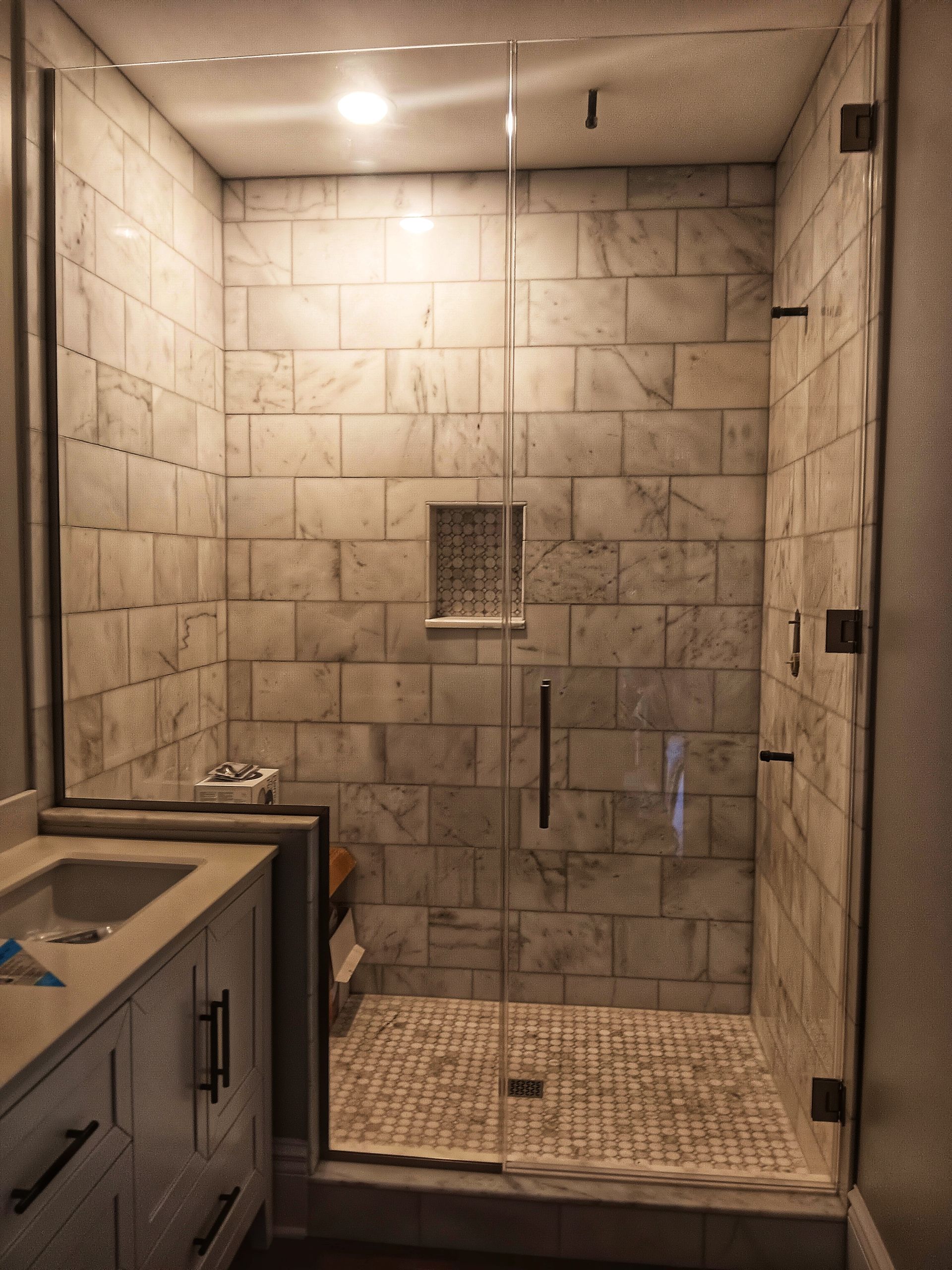 Bathroom shower with marble-like tile walls and a glass door. A small decorative niche and a gray stone floor are visible.