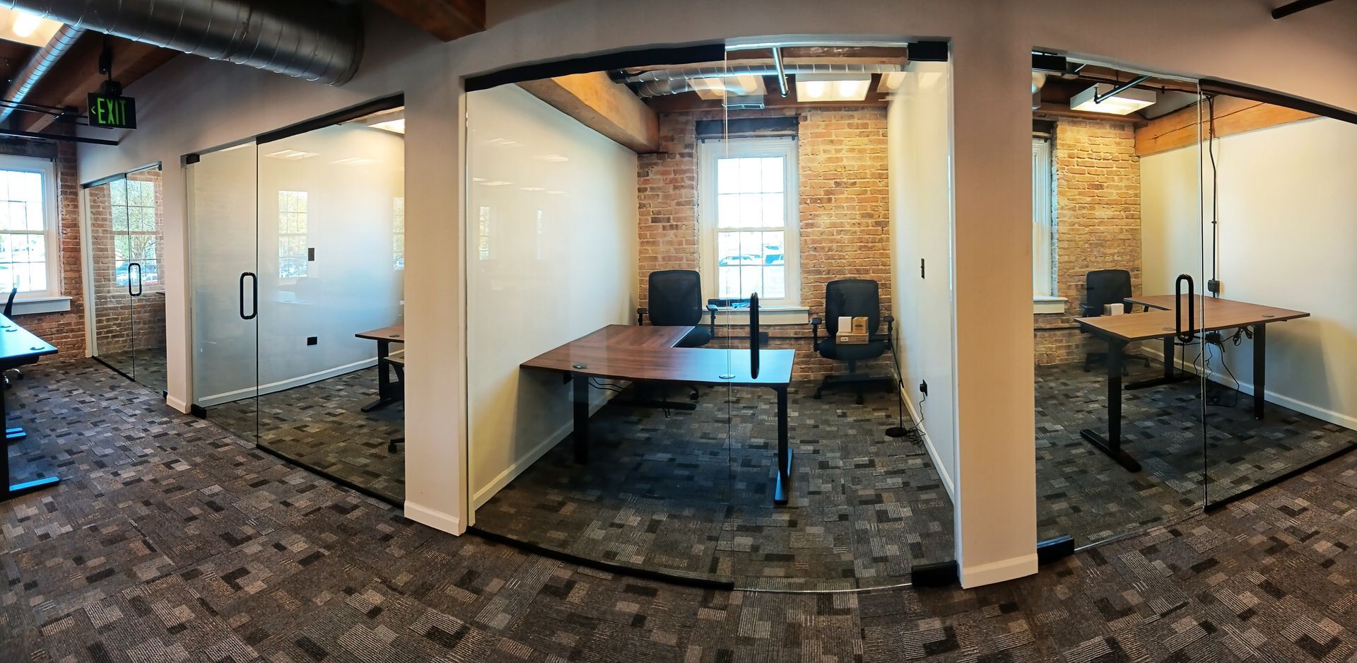 Office with glass-walled rooms, exposed brick, and desks, lit by natural and overhead lights.