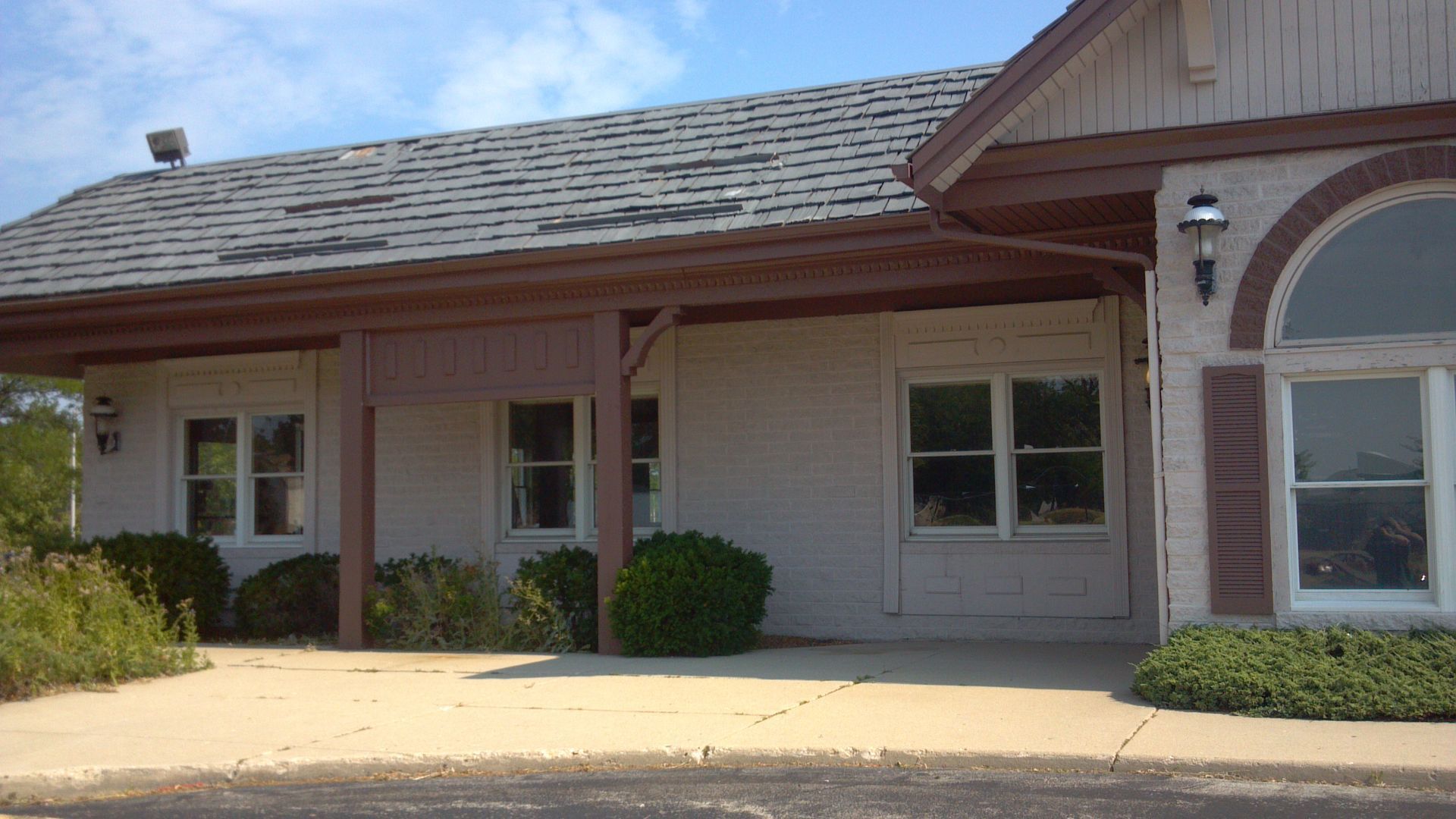 Cream-colored building with a porch and arched window. Gray roof, brown trim, and bushes in front.