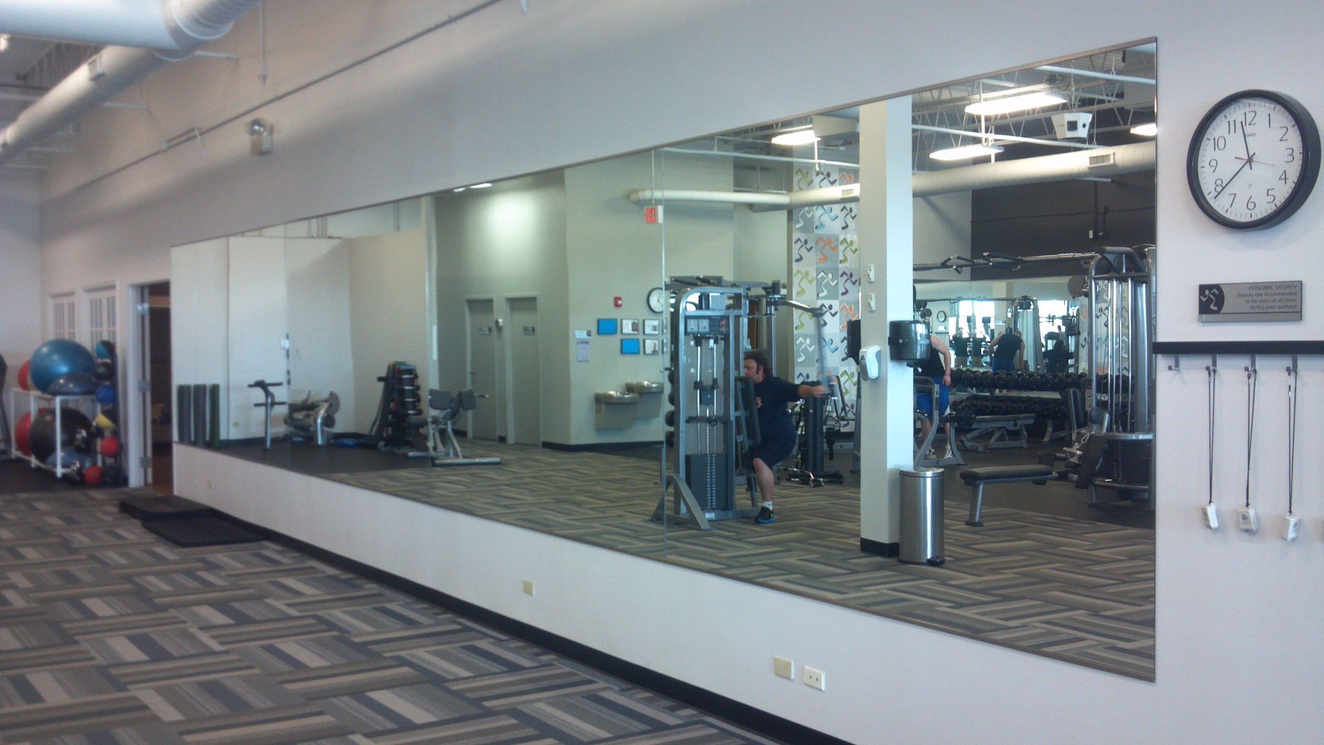 Gym with large mirror; people exercising. Gray carpet, white walls. Clock and equipment visible.