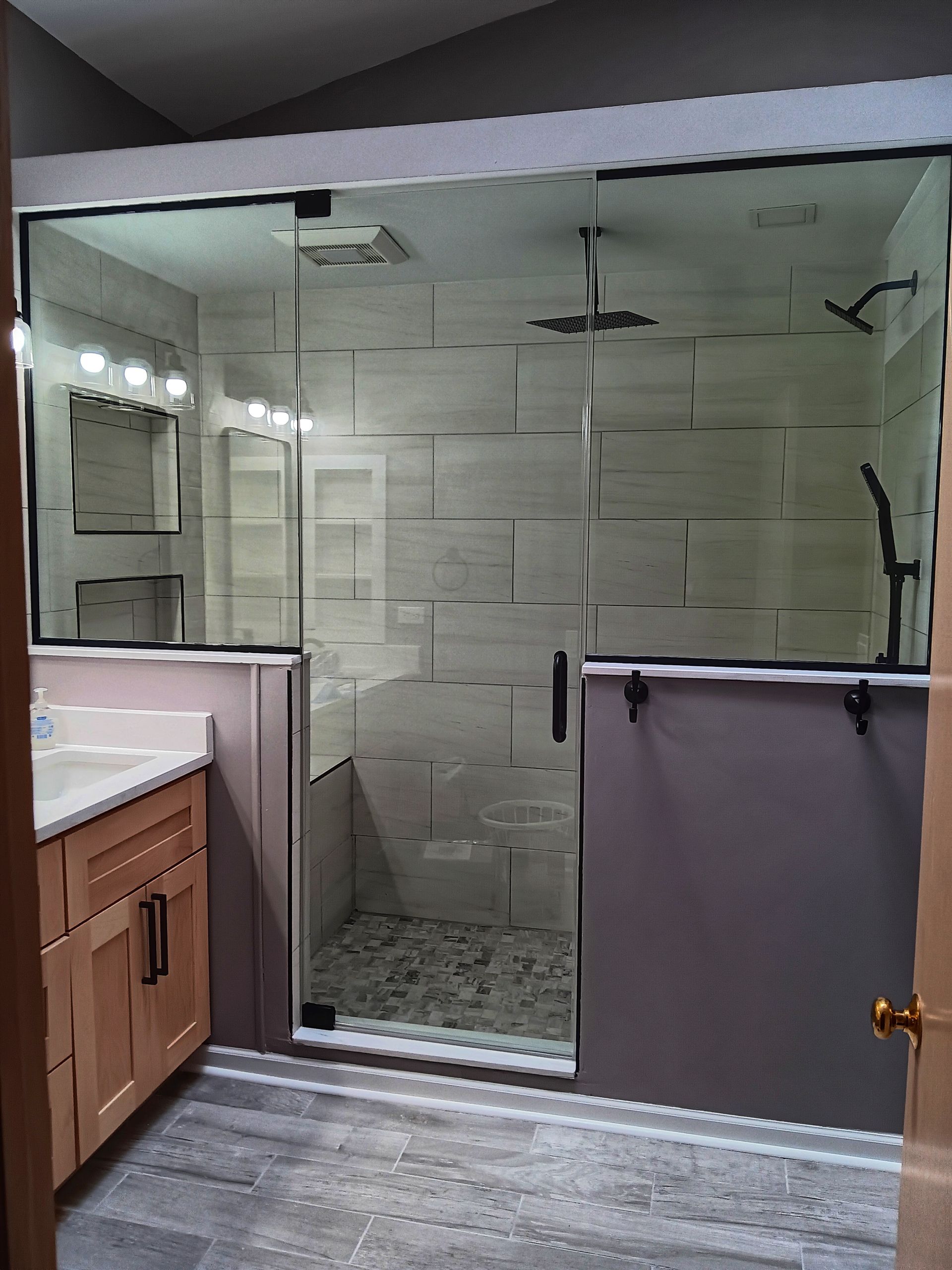 Bathroom with glass shower doors, stone tile walls, light wood vanity, and gray flooring.