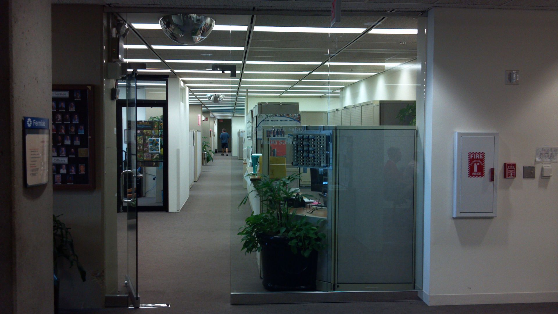 Glass doors open to a long office hallway lined with cubicles. A plant and fire alarm are visible.