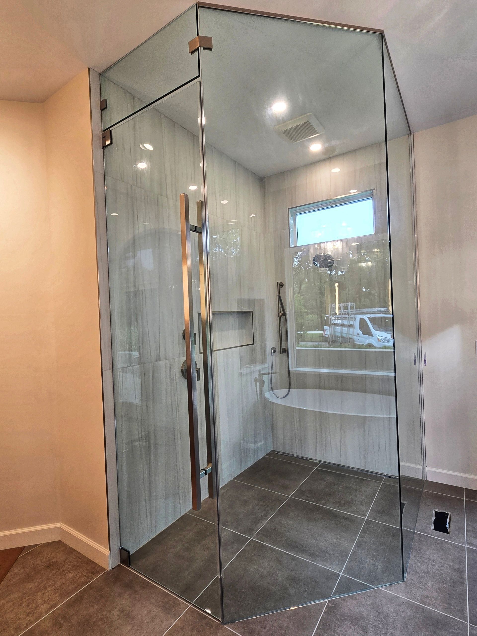 Glass shower enclosure in a bathroom with gray tile, bench, and window.