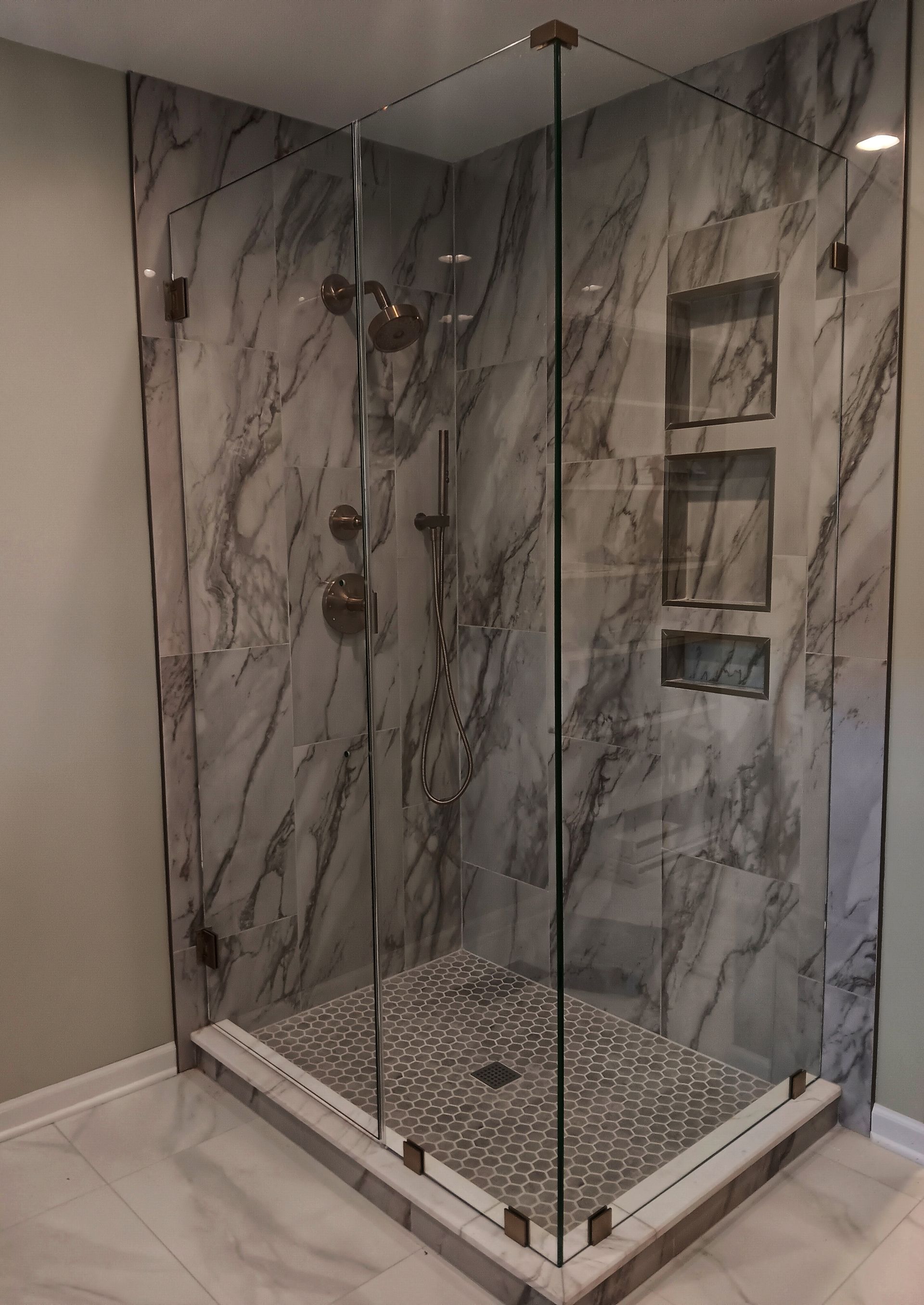 Glass-enclosed shower with marble-look tile walls, built-in shelves, and a pebble tile floor.