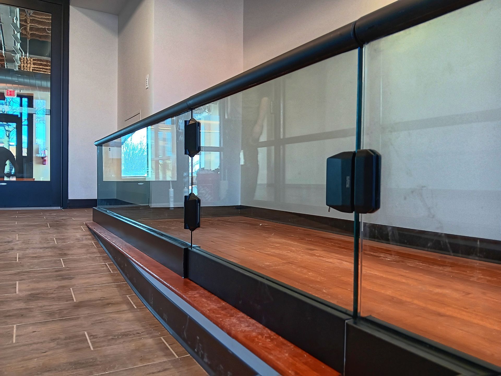 Glass railing with black top rail and posts, brown wood bench and flooring. Building interior.