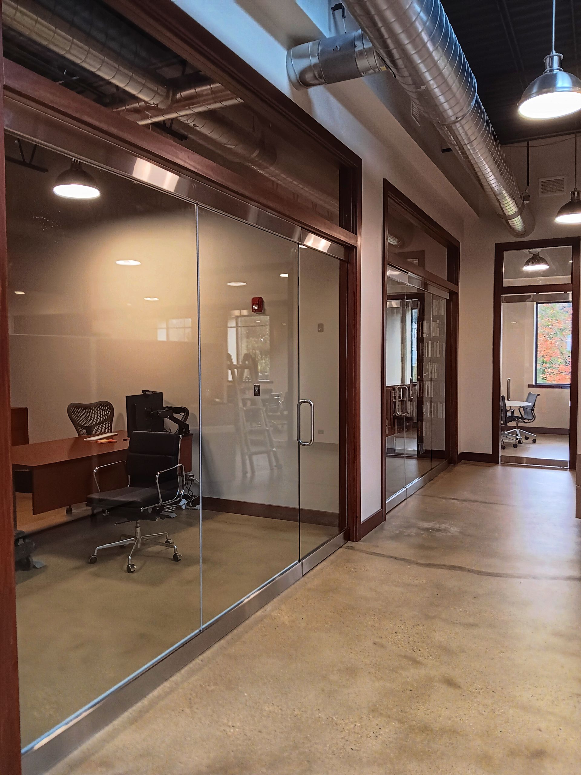 Office hallway with glass-walled rooms, wood trim, concrete floor, overhead lighting.