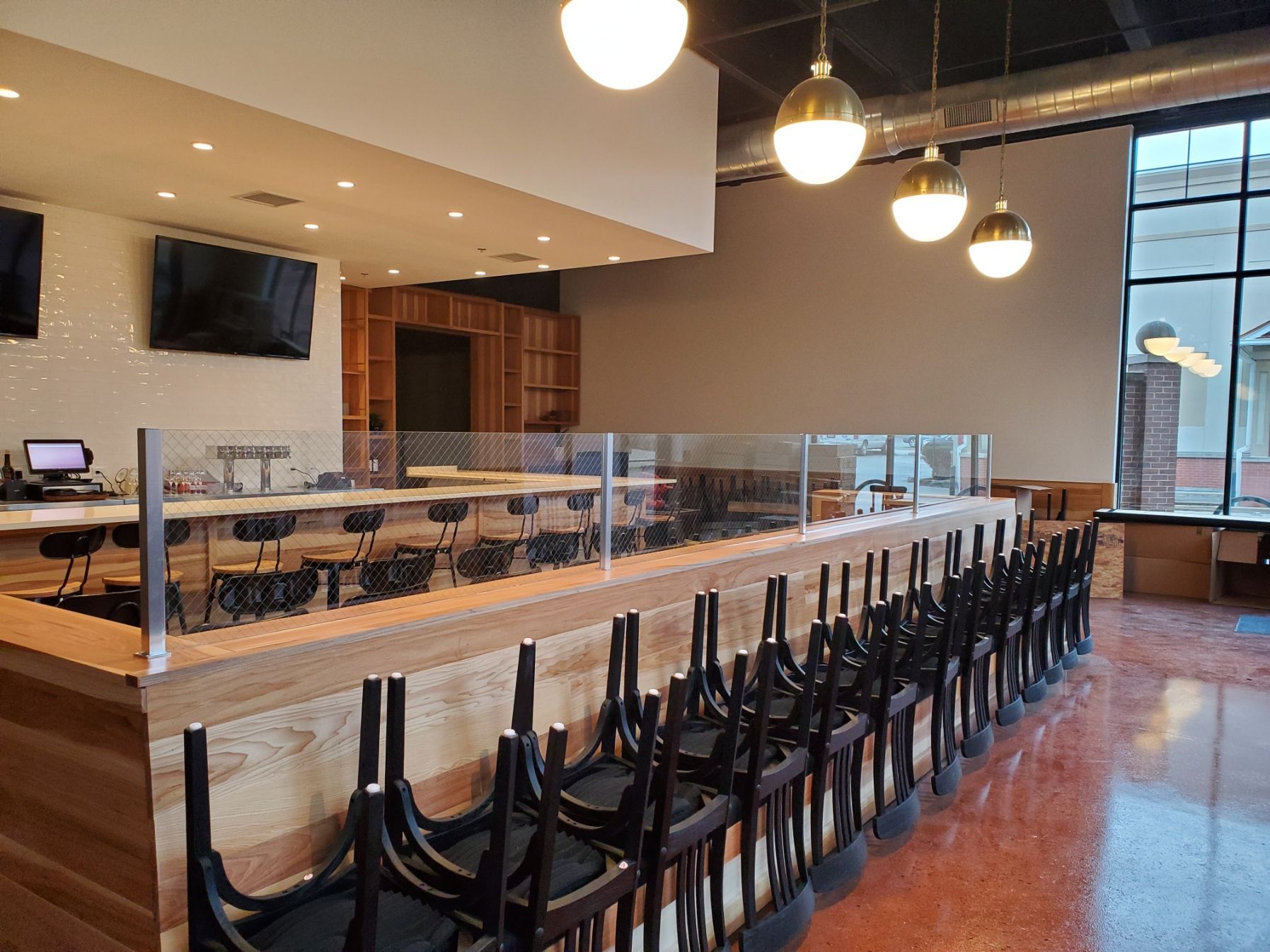 Empty bar area with closed chairs, clear partition, wood bar, and hanging lights.