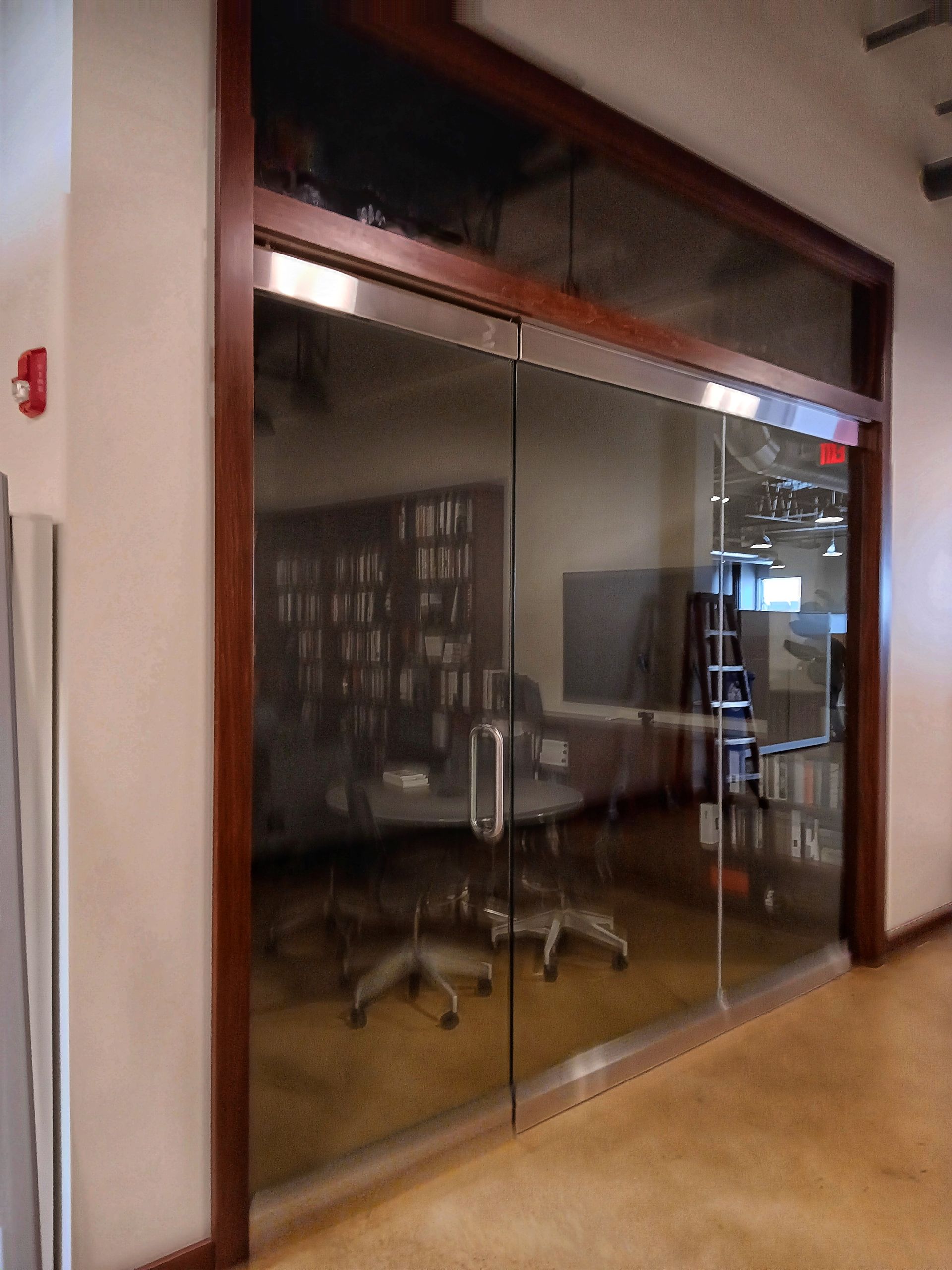 Glass-walled office with brown trim and stainless steel hardware, reflecting bookshelves and a desk.