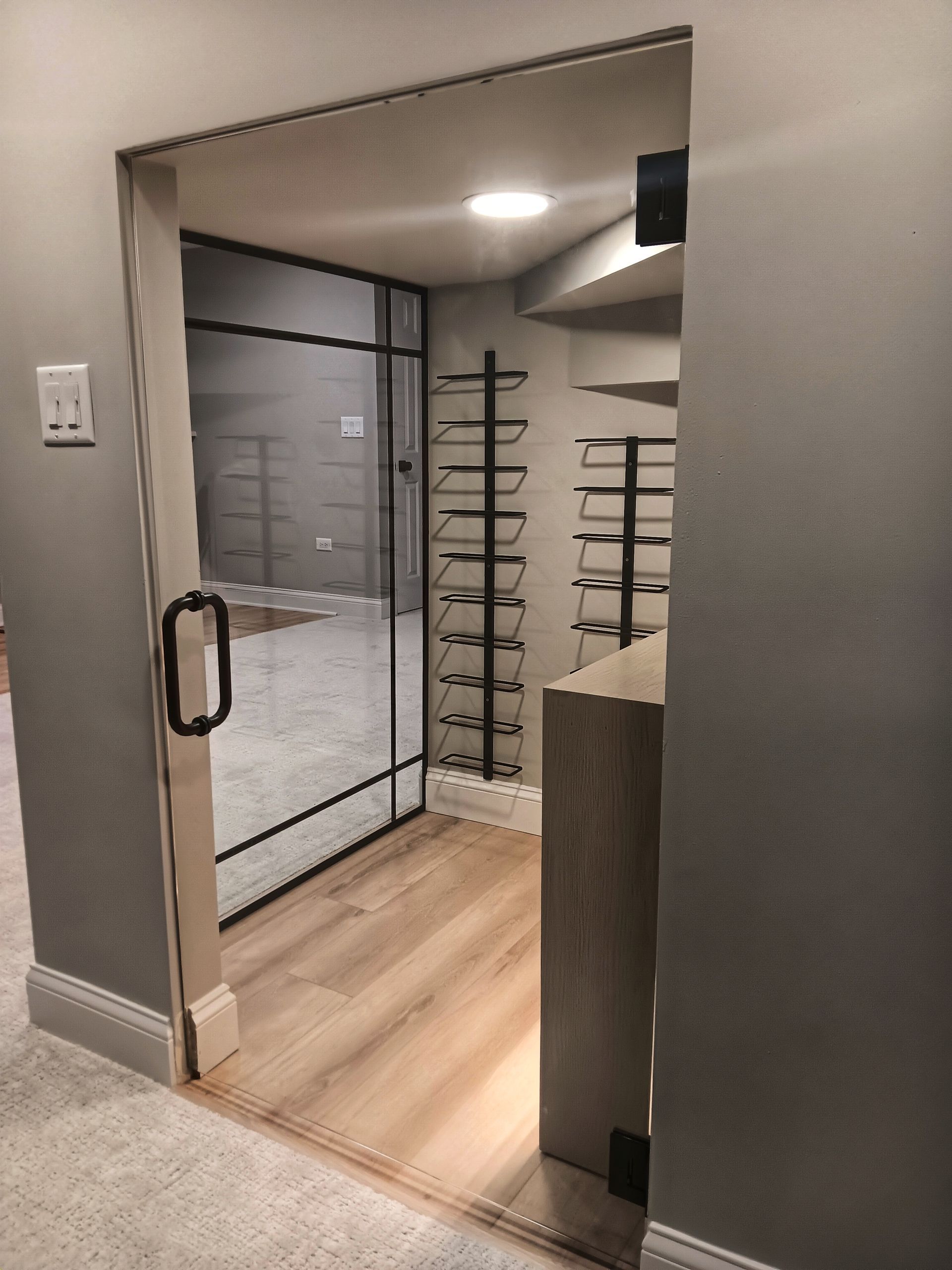 Glass-enclosed room with black trim, holding shelves and door. Light wood floor, gray walls.