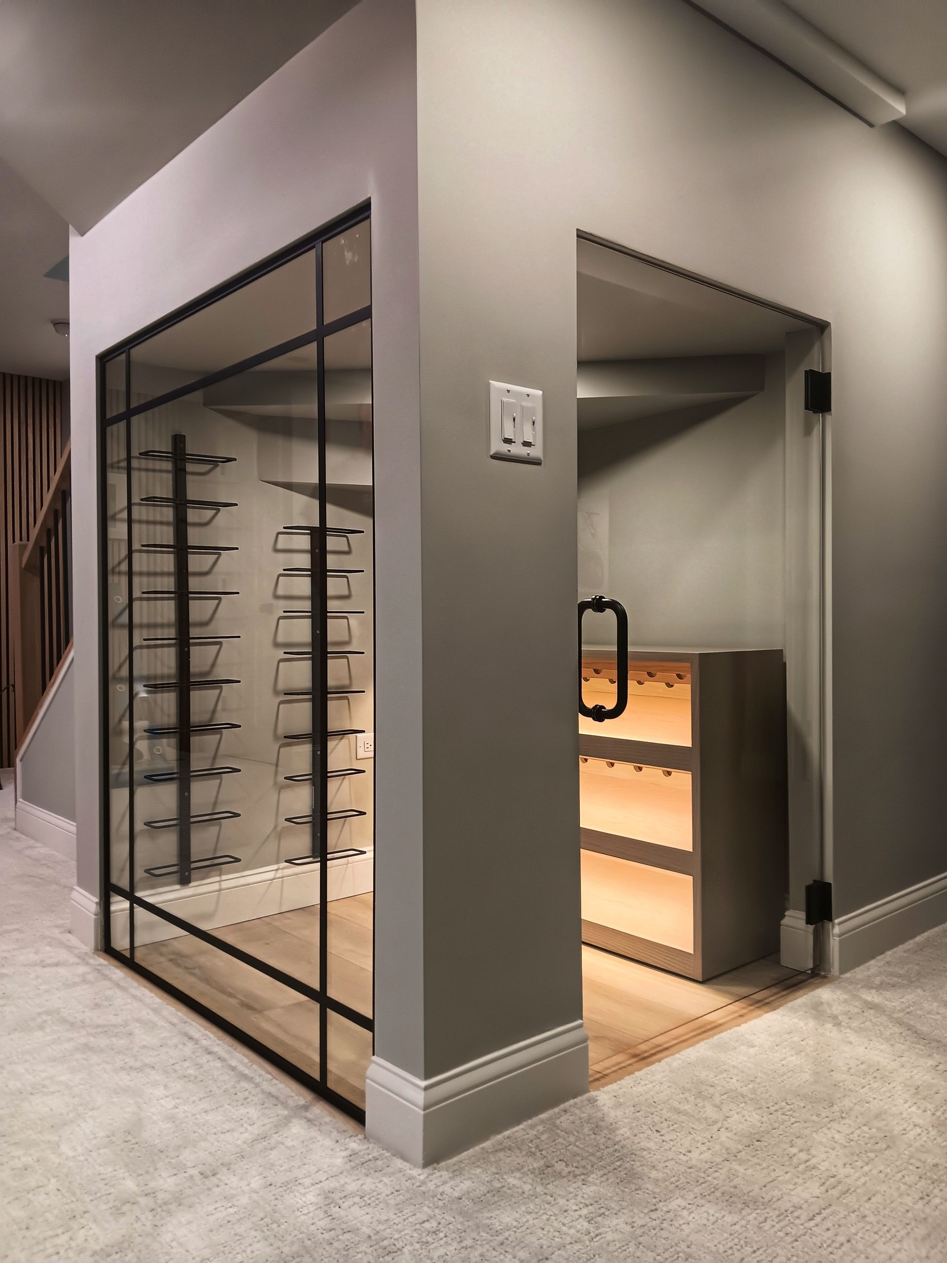 Wine cellar with glass-fronted display and door, inside a larger room with light gray walls and carpet.