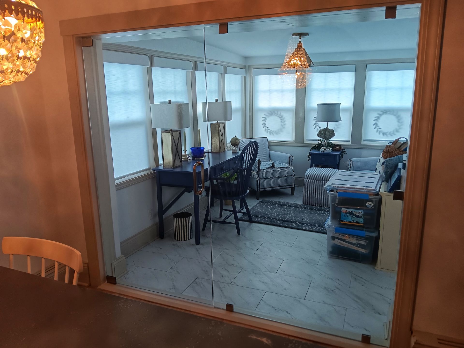 A sunroom with glass doors, furnished with desk, chairs, sofa, and lamps, lit by natural and artificial light.