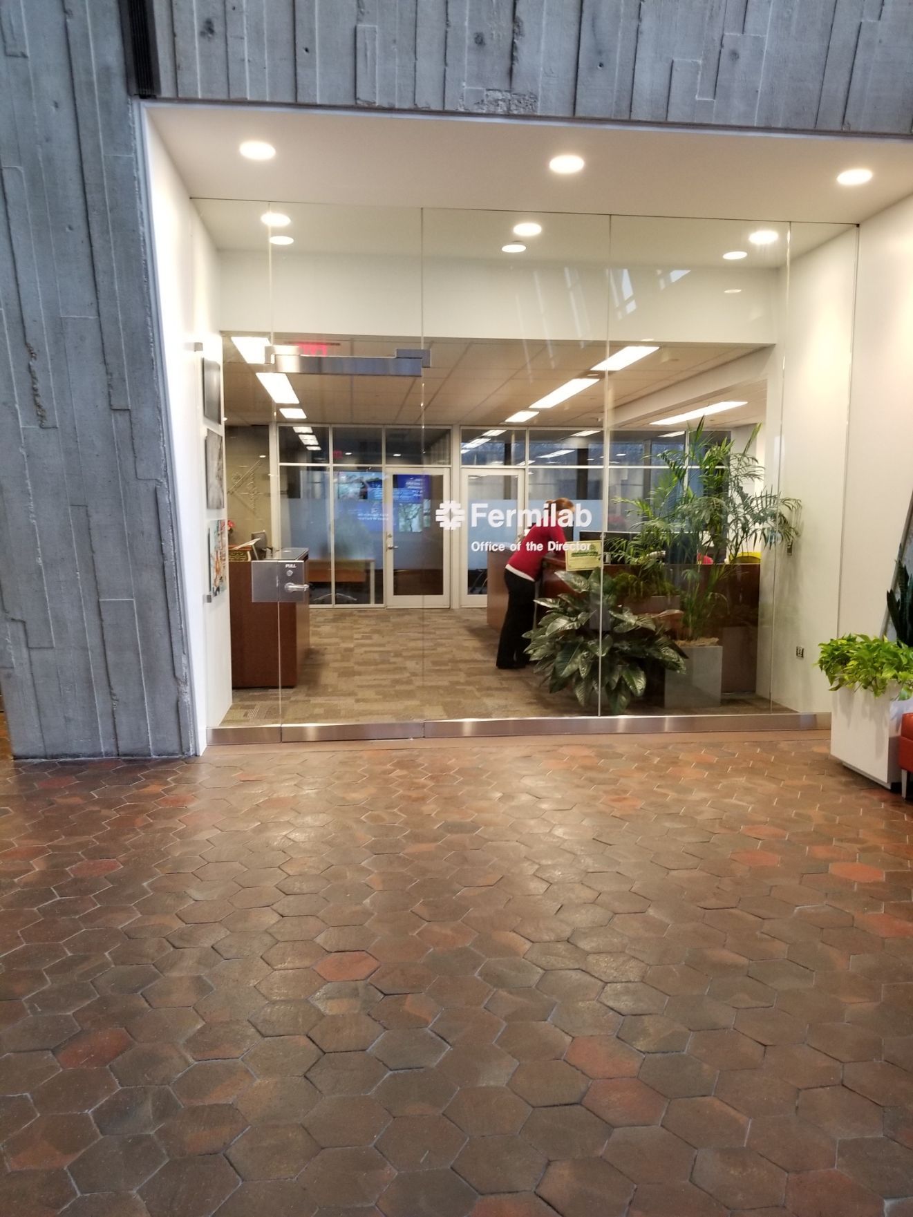 Entrance to the Fermilab with glass doors, a person, and plants. Brown brick flooring and concrete walls.