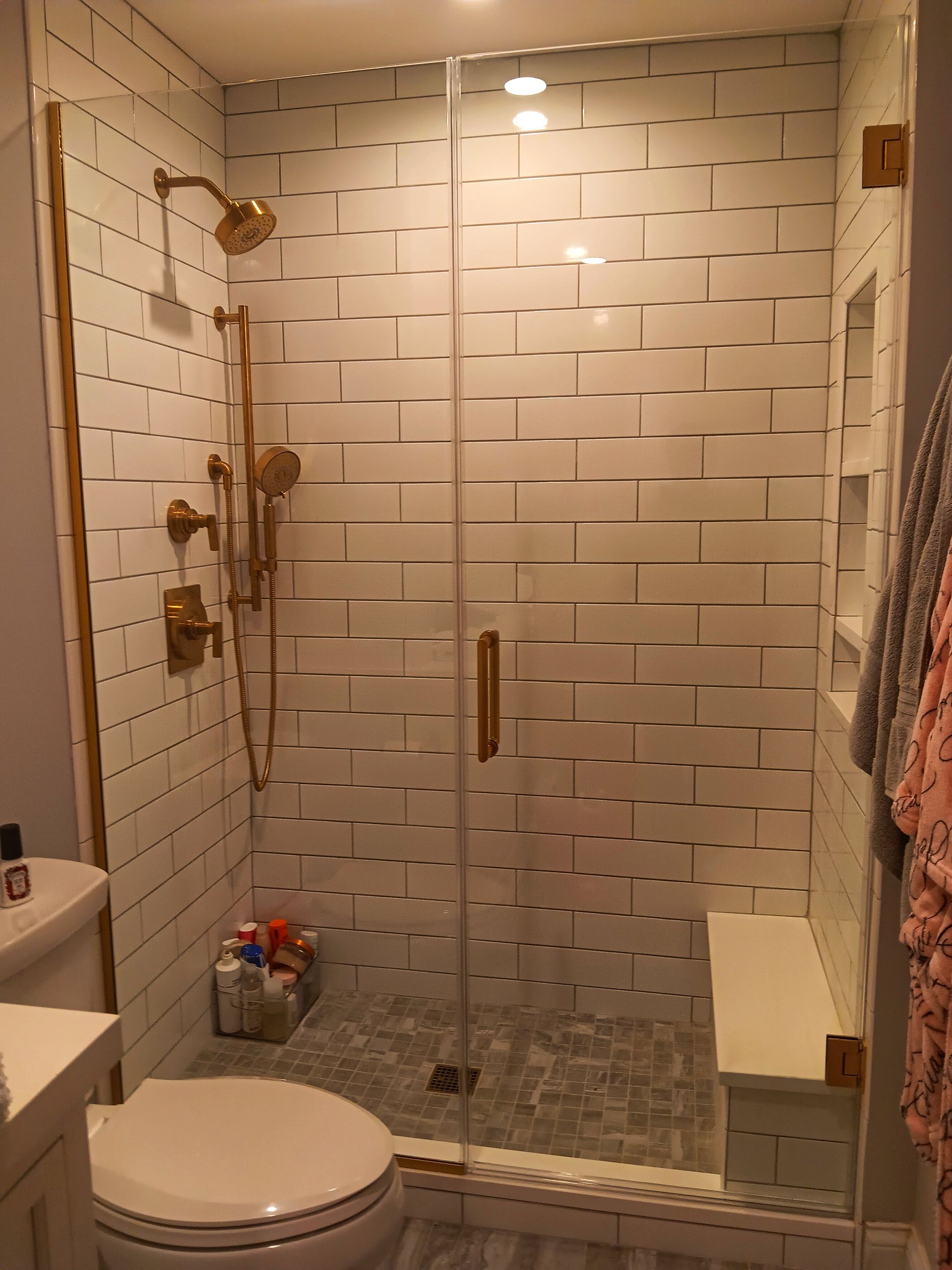 A small, modern bathroom with gold fixtures. Shower has a bench and white brick-like tiles. Toilet is in the foreground.
