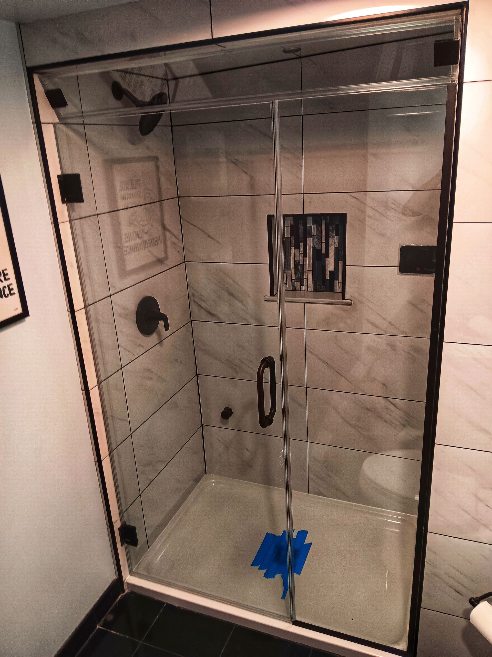 Glass shower enclosure with black fixtures and marble-look tile walls. Blue tape on the shower floor.