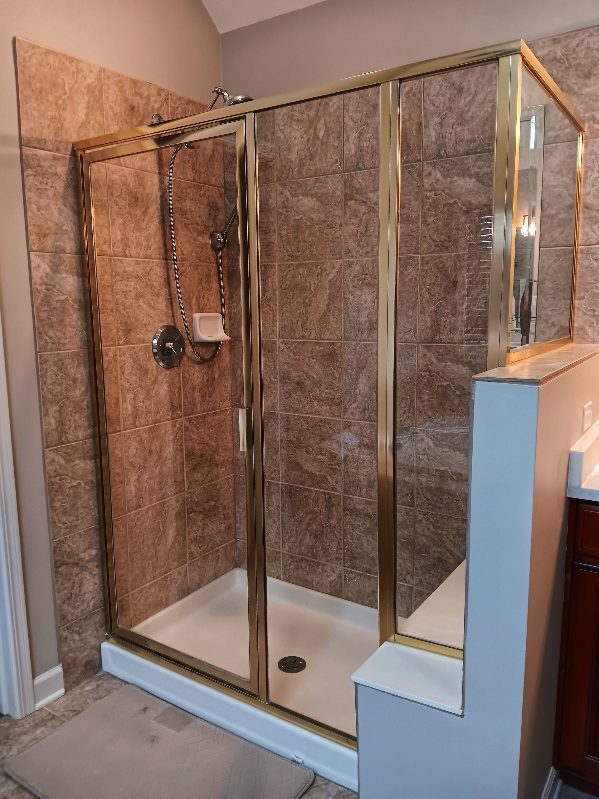 Gold-framed shower with textured brown tiles. White shower base and small ledge.
