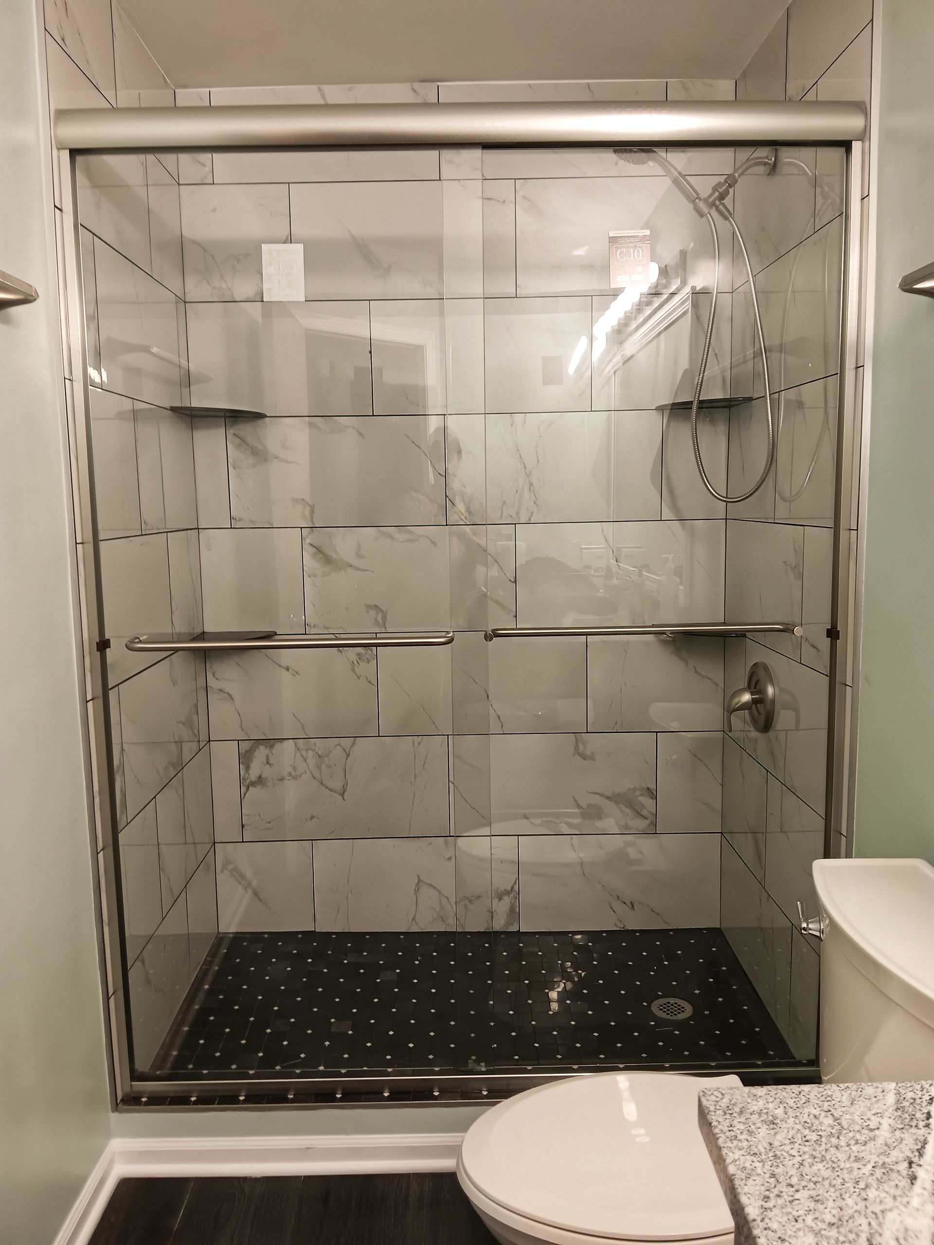Shower with glass doors and marble-like tile walls. The shower has shelves and a dark floor. A toilet is next to it.