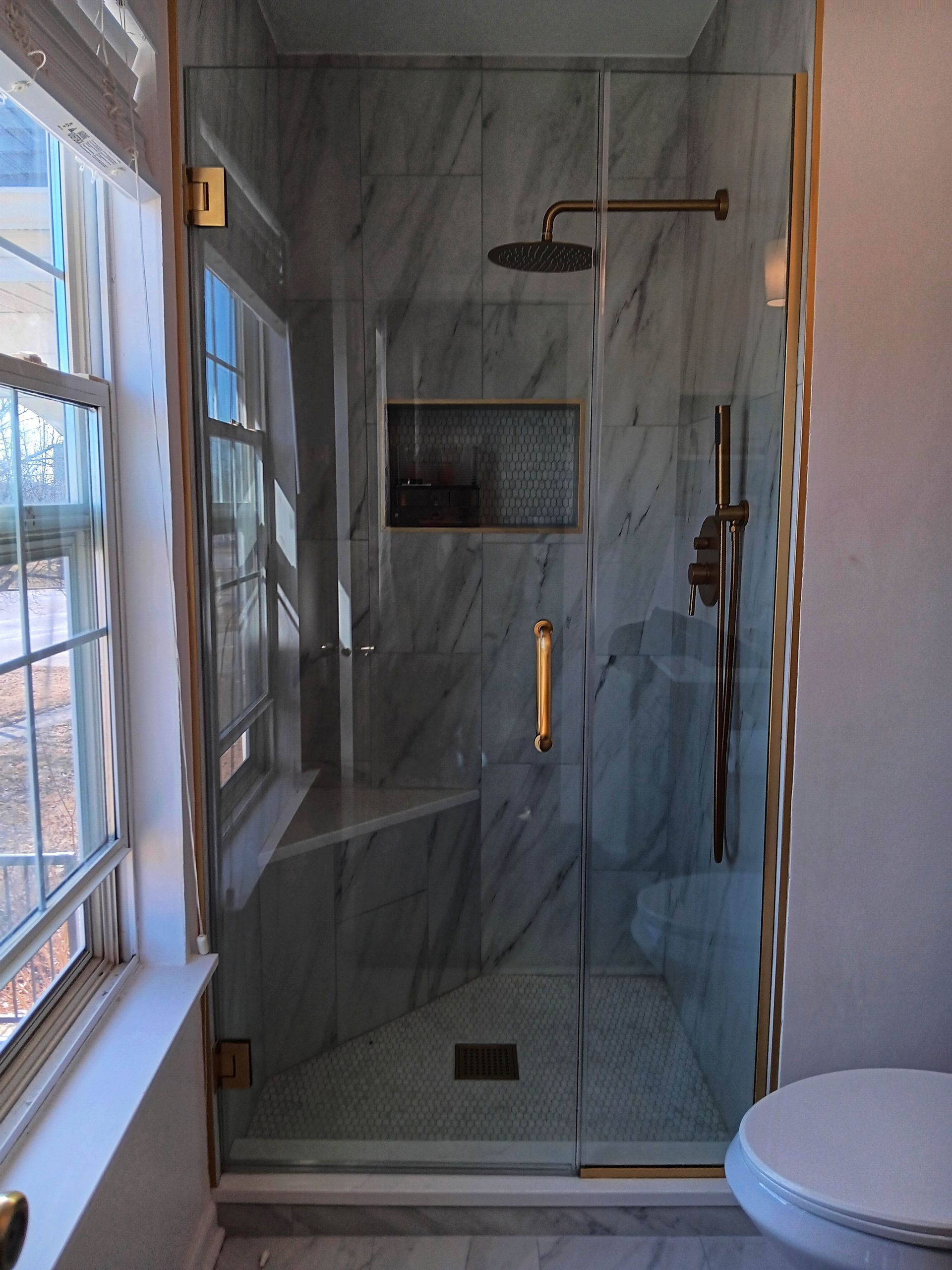 Glass shower with gold trim and fixtures, marble-look tile, and a window.